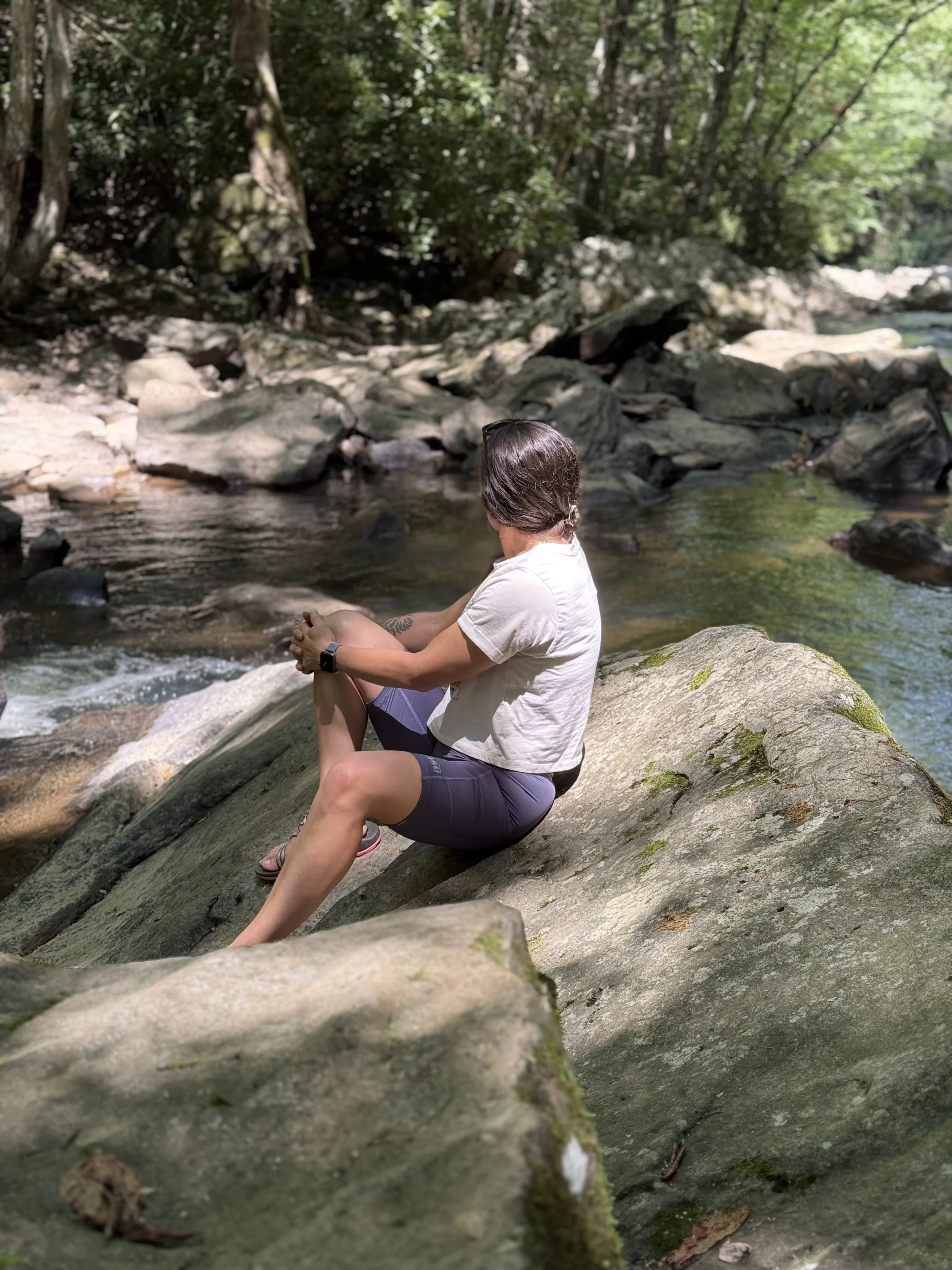 Person sitting on a large mossy rock by a shallow stream in a forest, looking away from the camera with trees and rocks in the background.