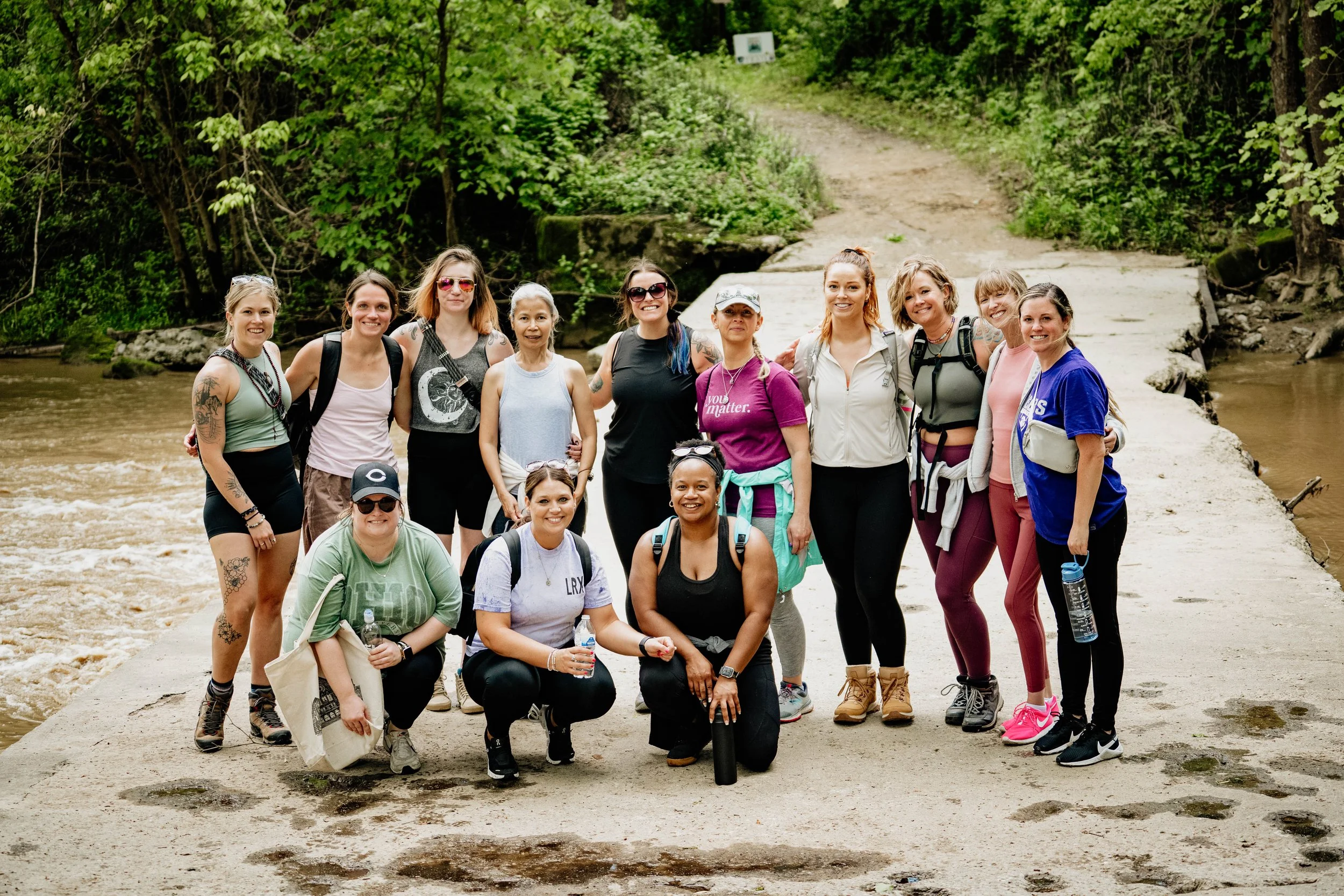 Group of women hikers standing on a trail near a river in a forested area, smiling at the camera.