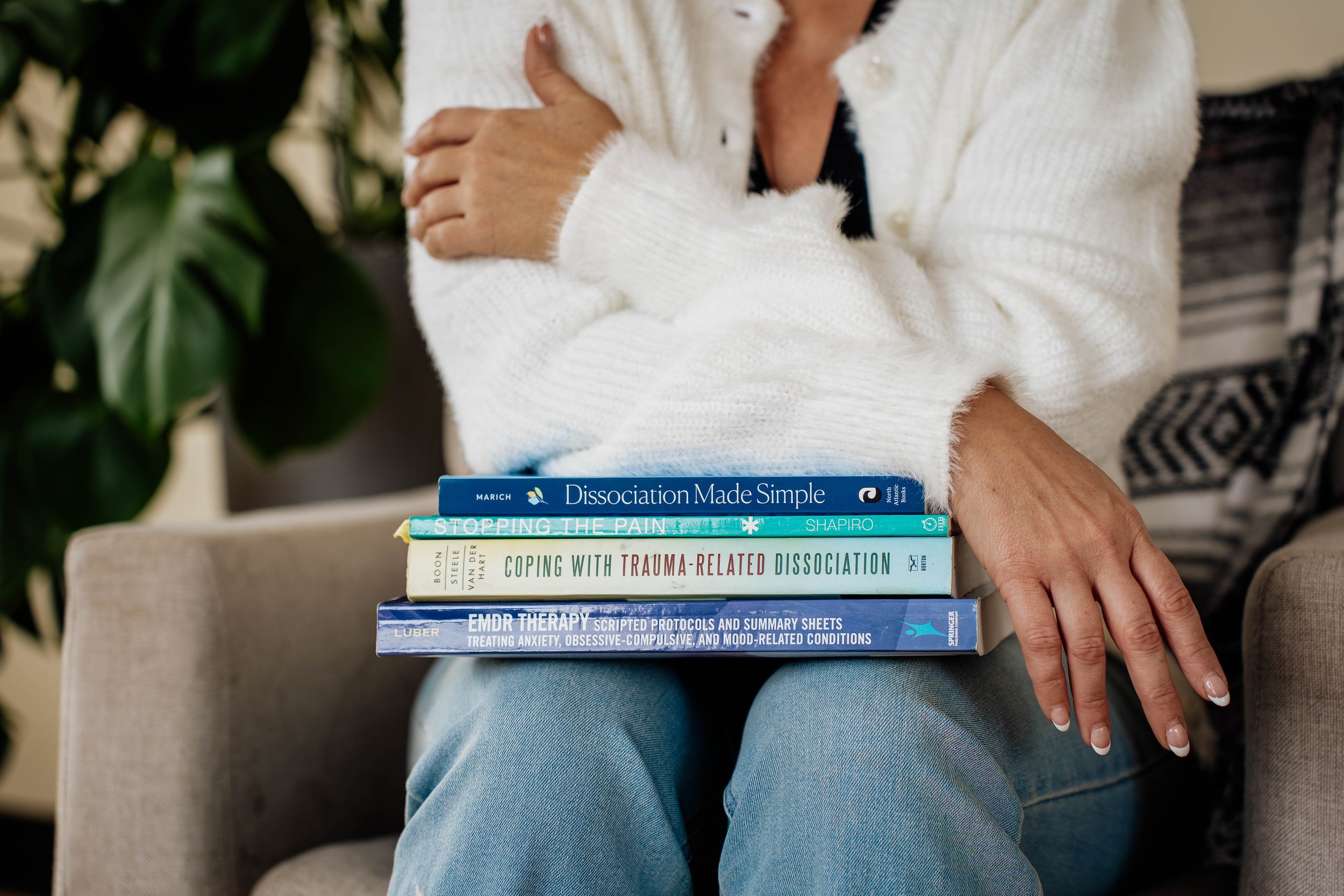A person wearing a white sweater and blue jeans sitting on a beige chair with a stack of books on their lap.