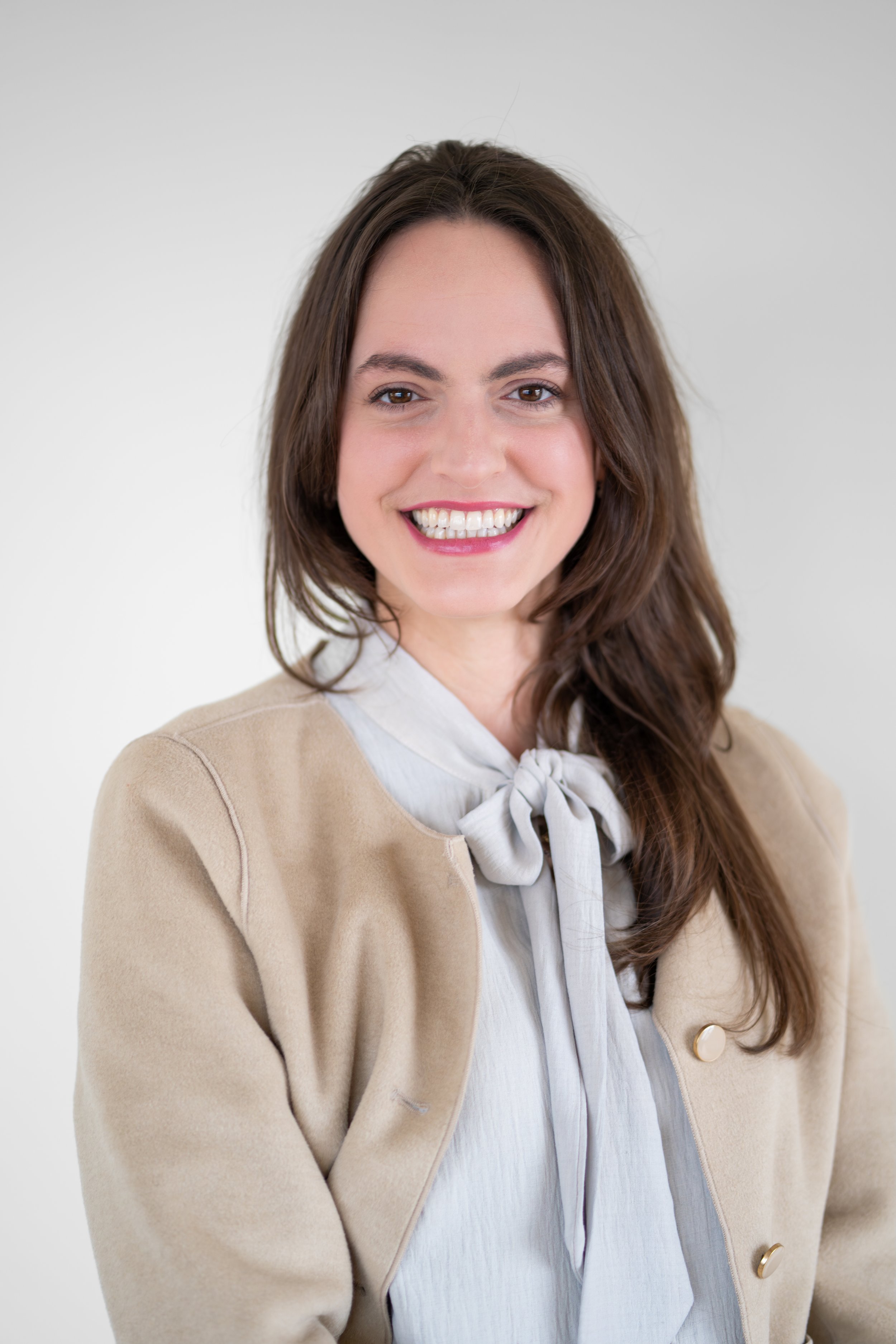 A woman with long brown hair, smiling, wearing a beige blazer over a white blouse with a bow, standing against a plain white background.