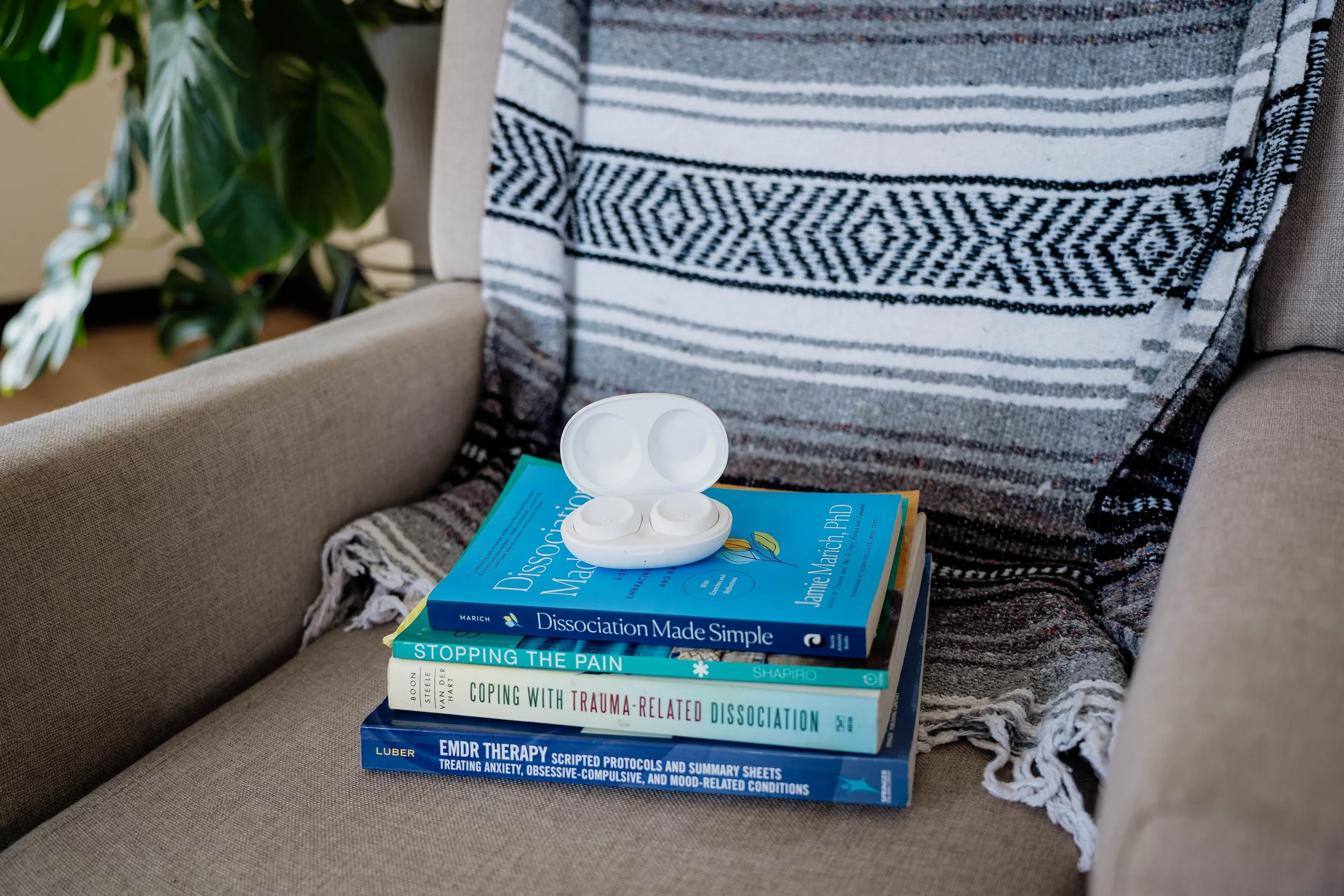 A stack of six psychology and therapy books on a beige couch, with a white open case of wireless earbuds on top, a patterned throw blanket, a plant with large green leaves, and part of a striped pillow in the background.