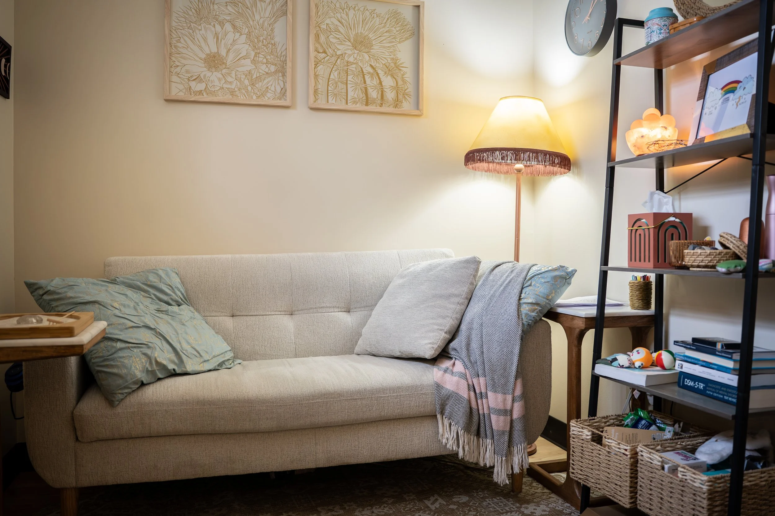 A cozy living room corner with a beige sofa, light green pillows, a wooden side table, a tall standing lamp with a fringed shade, and a black shelving unit with various decor and books. Three framed floral line art prints hang on the pale yellow wall above the sofa.
