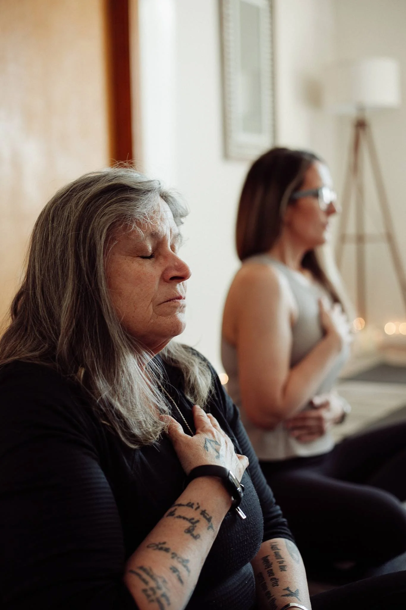 Two women sitting on a bench with eyes closed, possibly meditating, in a room with a mirror and a standing lamp in the background.