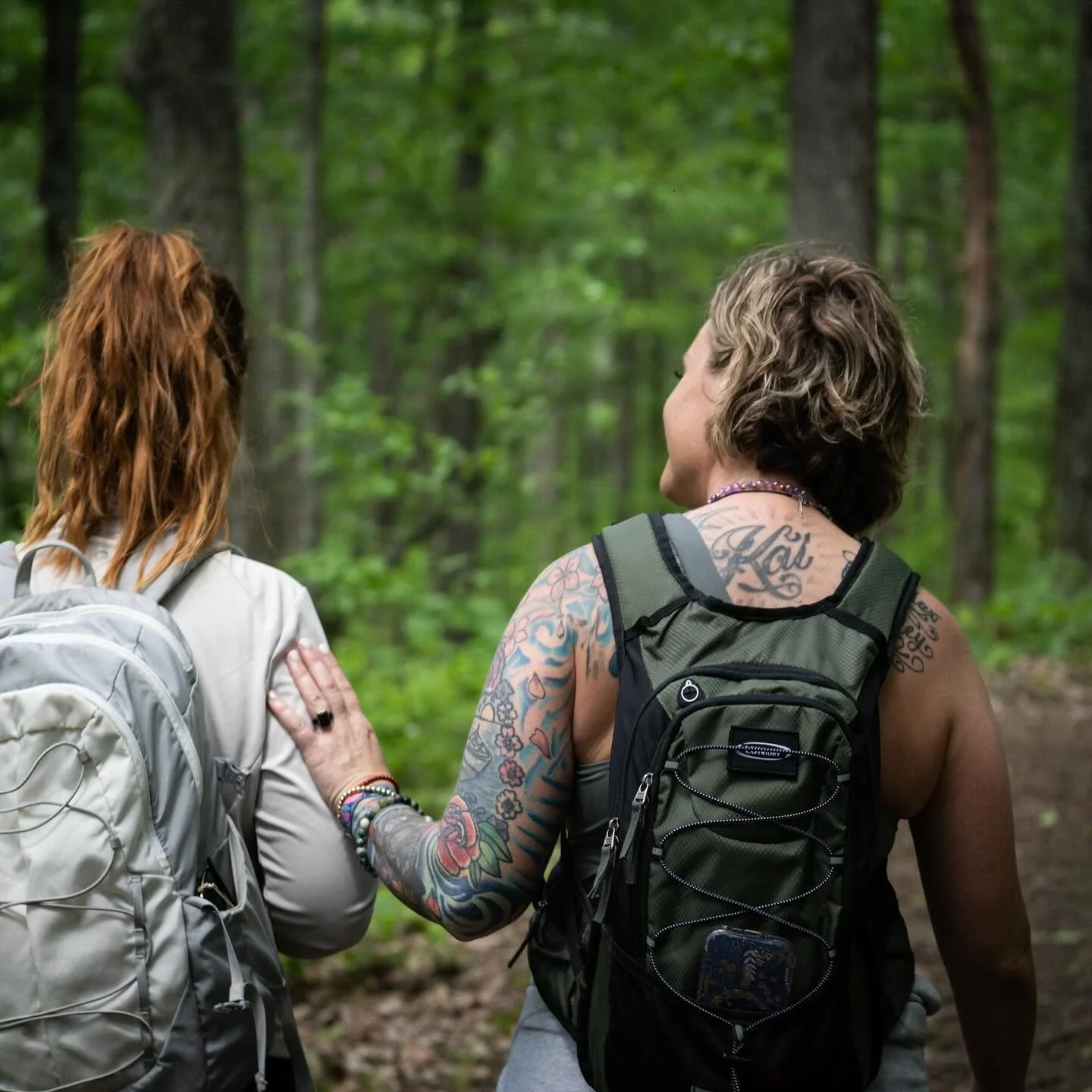 Two women wearing backpacks walking in a green forest, with one woman placing her hand on the other's shoulder.