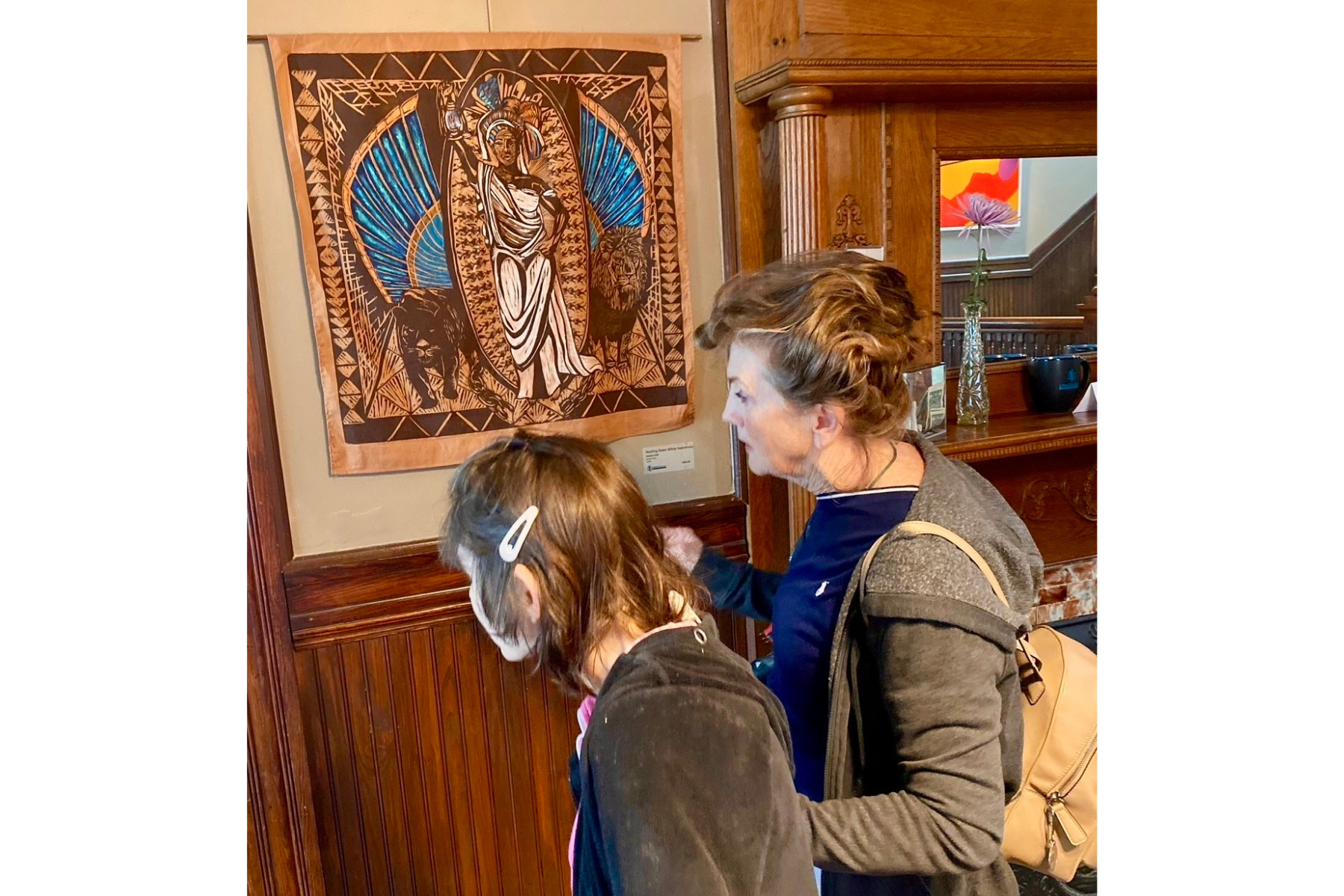 A woman with short curly hair and a beige purse looks at artwork on a wall with a young girl with glasses and long brown hair. The artwork is a colorful African-style painting with a woman, lions, and animals.