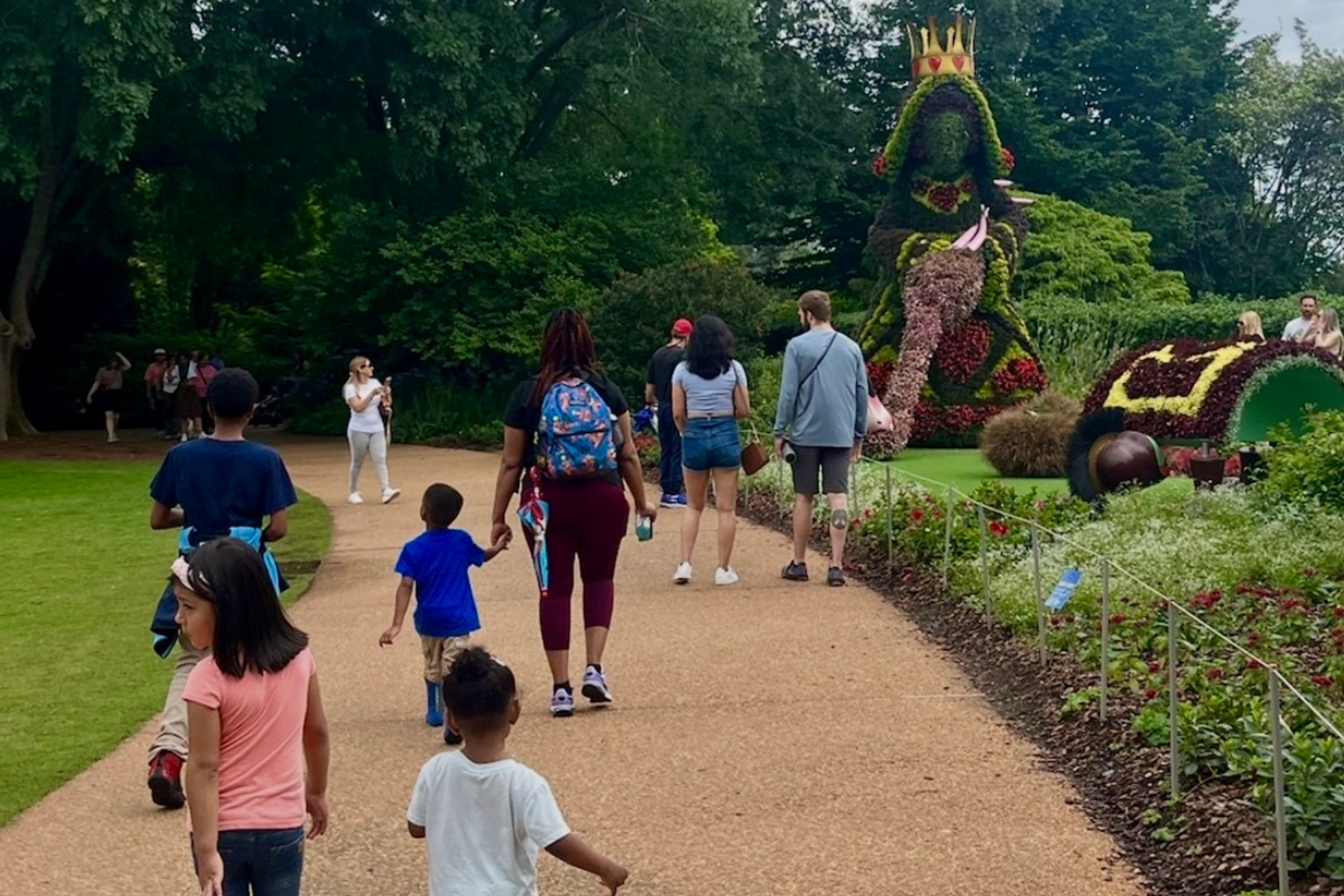People walking along a pathway in a garden with large Alice and the Wonderland floral sculptures, including a crown and a floral vehicle, surrounded by lush green trees and plants.