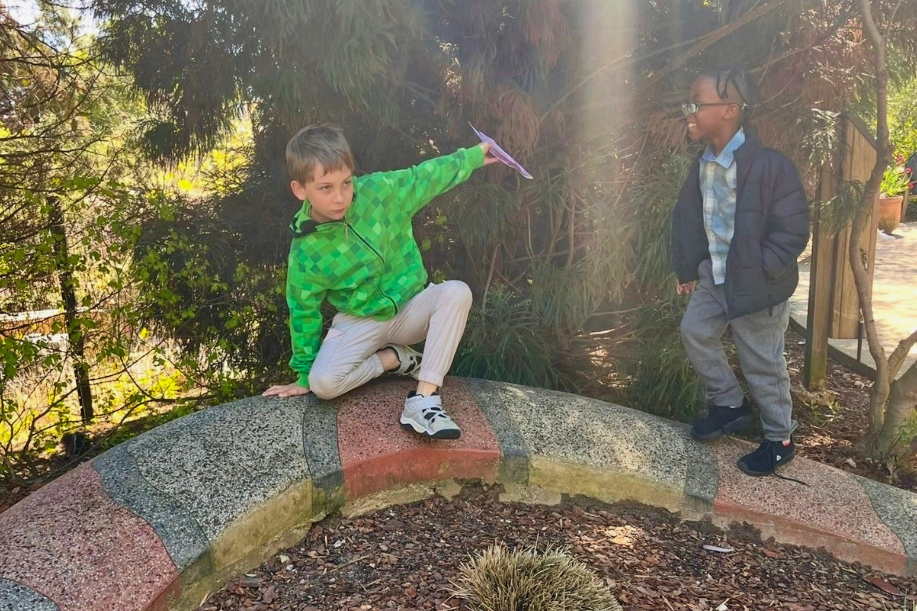 Two boys outdoors near a curved stone garden border, one sitting on it and the other standing.