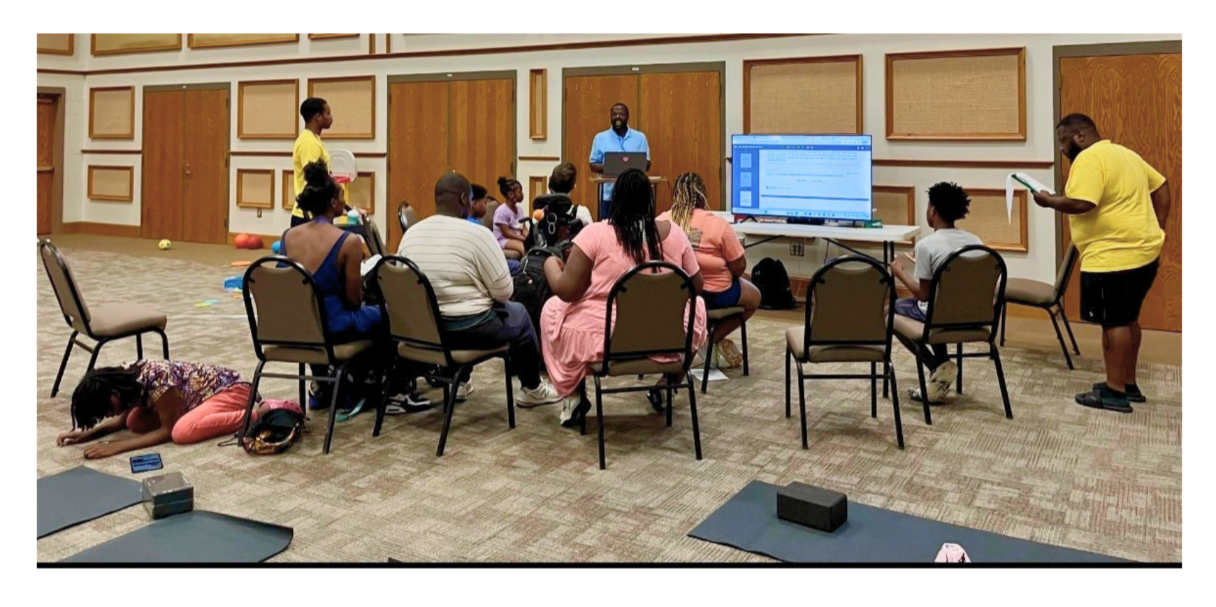 A diverse group of children and adults attending a class. The instructor stands at a podium with a laptop, speaking to the audience. Some attendees are sitting, some standing, and a child is lying on the floor.
