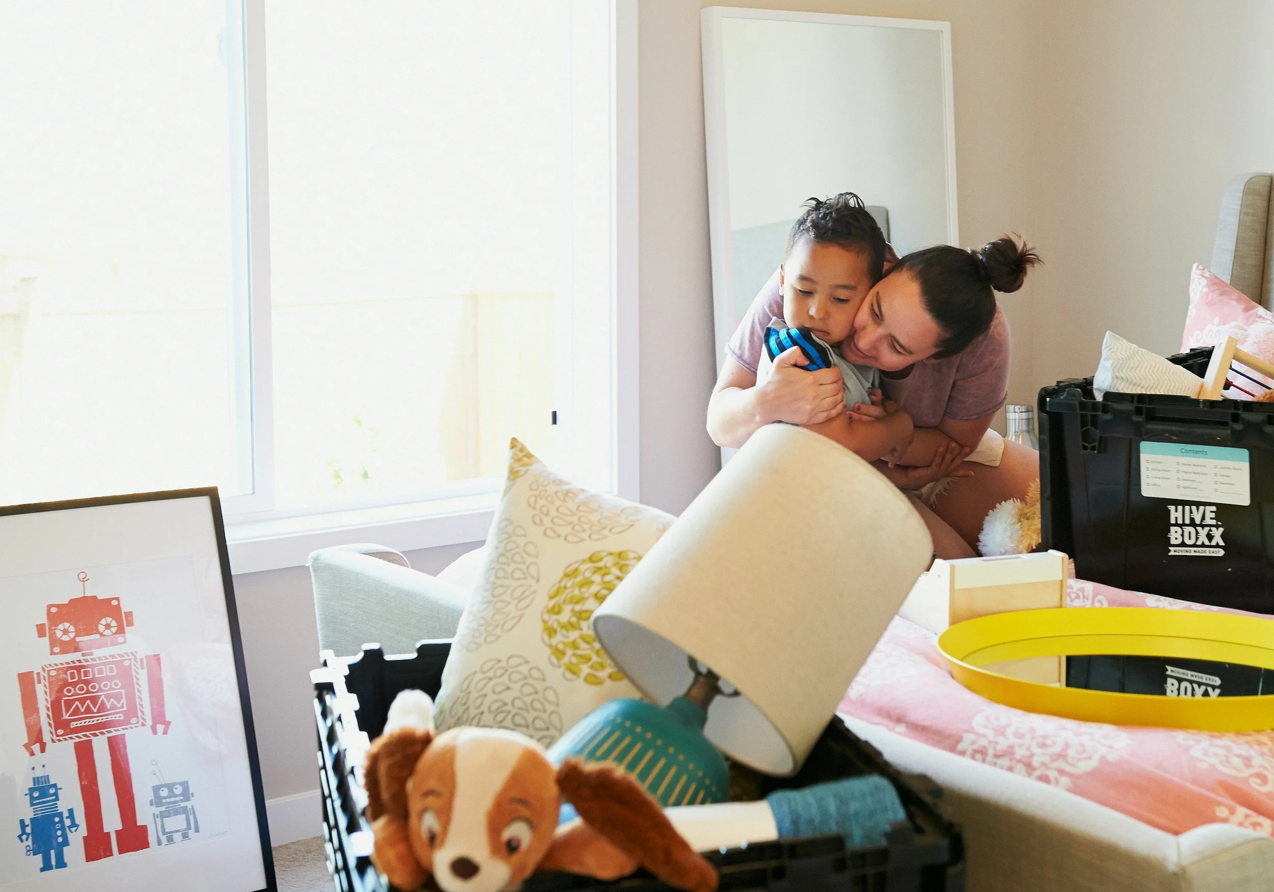 A woman hugging a young boy indoors near a large window. The scene includes household items and a table with various objects, with a whiteboard or picture frame visible in the background.