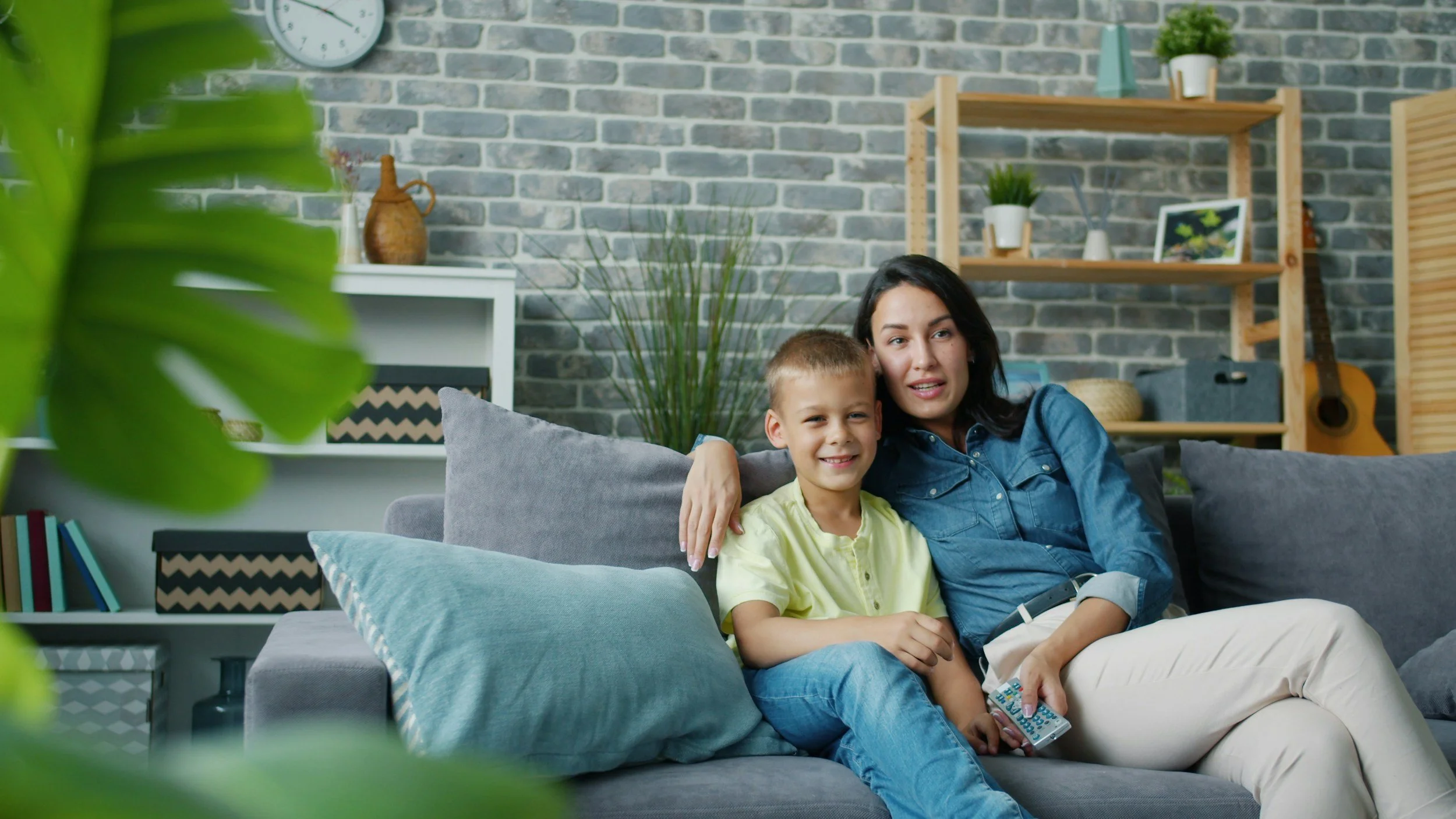 A woman and a son are spending time together sitting on a gray sofa in a modern living room, smiling and relaxing. The living room has a brick wall, shelves with decorative items, a guitar, and indoor plants.