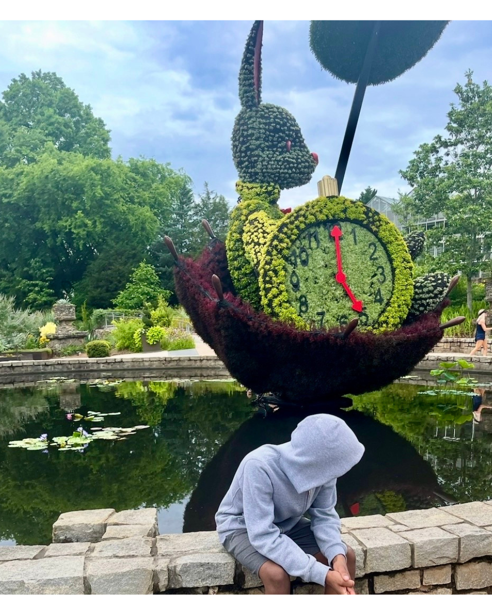 A person in a gray hoodie is sitting on a stone ledge beside a pond with lily pads. In the background, a large Alice and the Wonderland topiary sculpture of a rabbit holding a clock, made of colorful flowers.