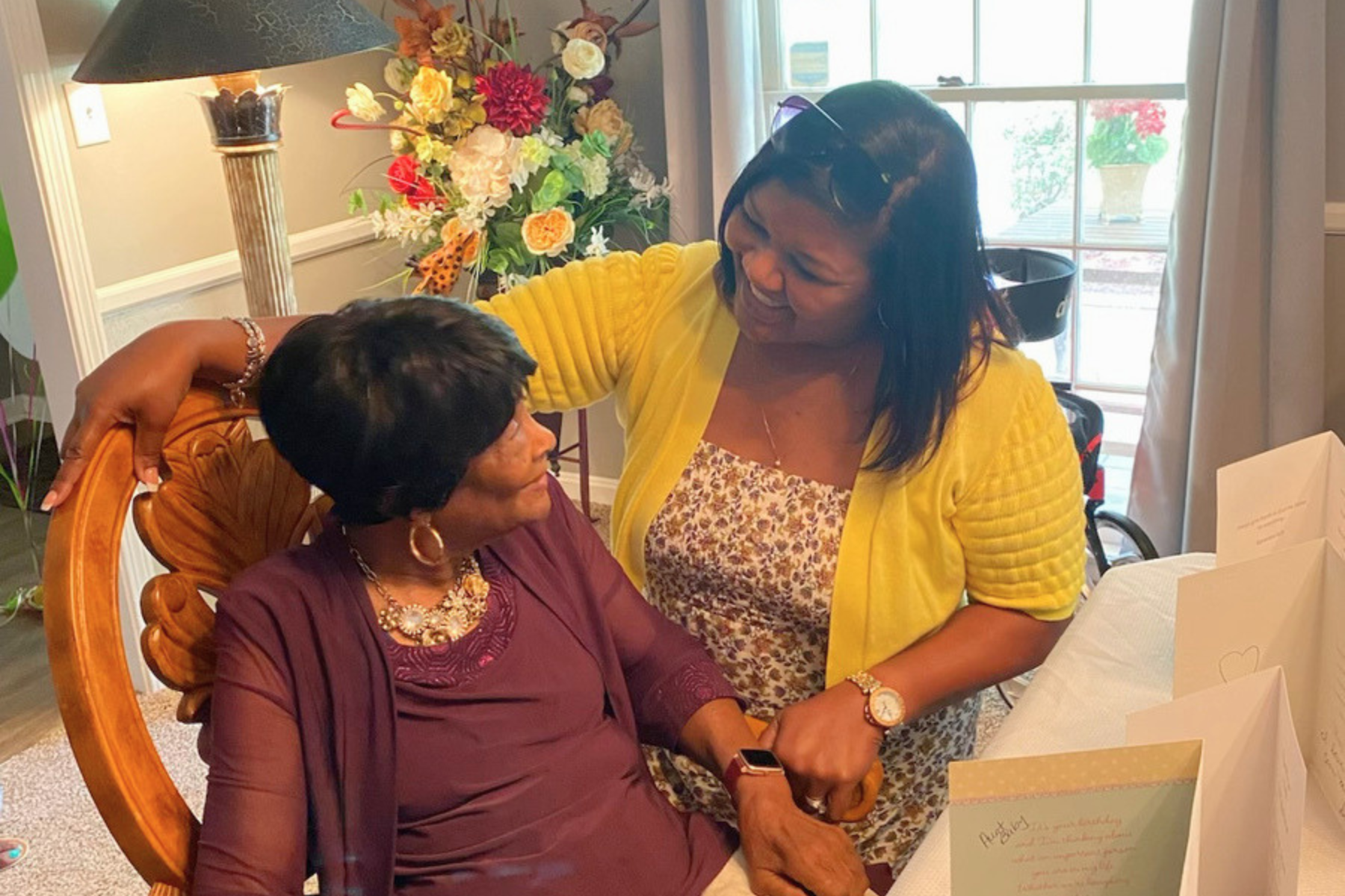 Two women sharing an emotional moment, one seated and the other standing, in a cozy, decorated room with flowers and windows in the background.
