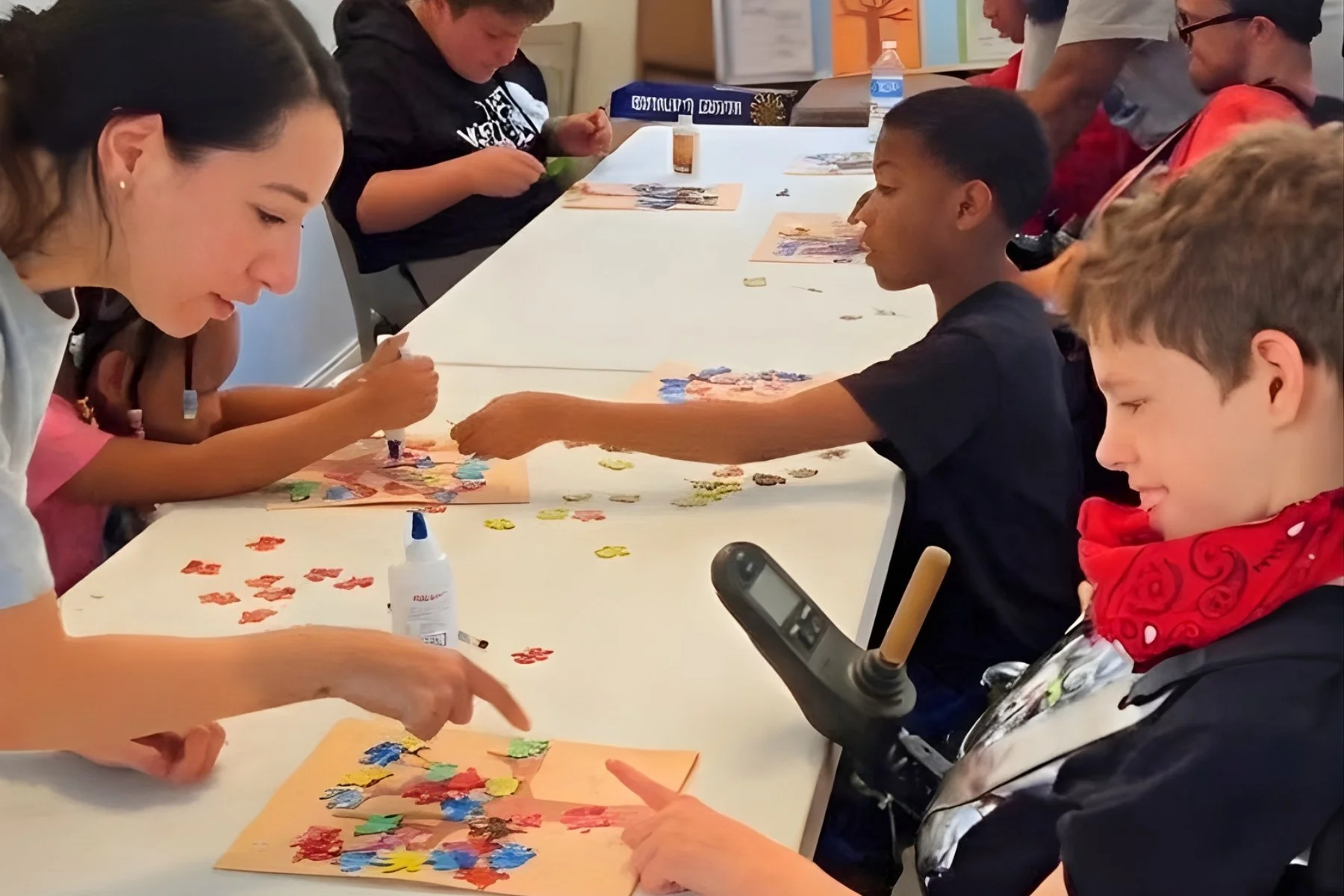 Participants of all abilities are engaging in conversation and creative activities together, sitting around a white table, creating artwork with colorful torn paper and glue, and participating in a craft activity.