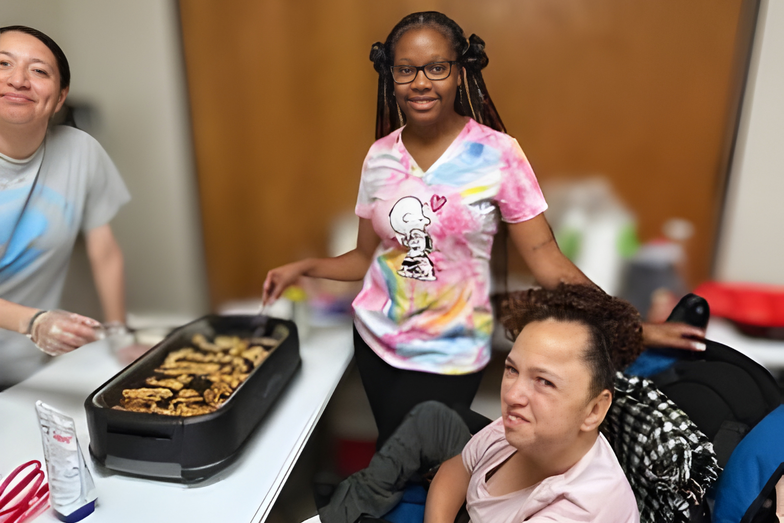 Three women in a kitchen preparing food, with a tray of cooked chicken on the table, one woman wearing glasses and a tie-dye shirt standing, the woman seated has curly hair, and the woman has short dark hair.