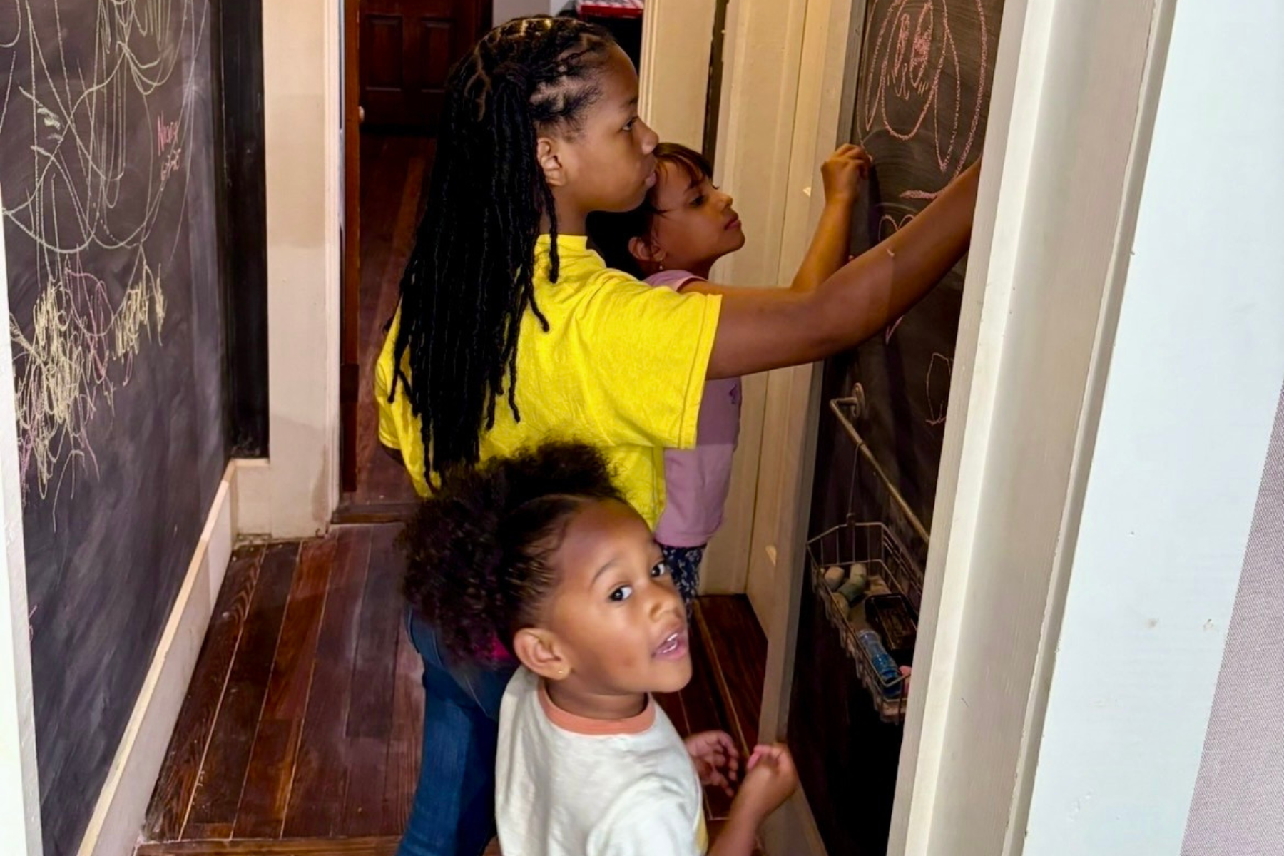 Three children in a hallway are drawing on a chalkboard with chalk while a fourth child looks at the camera.
