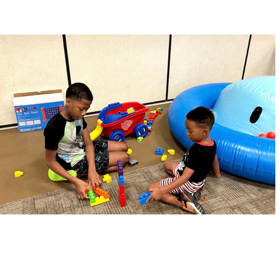 Children participate in imaginative play and hands-on learning activities together. Two young boys are playing with colorful building blocks on the floor in a play area with a large blue inflatable pool and a toy cart nearby, during Village Nights.