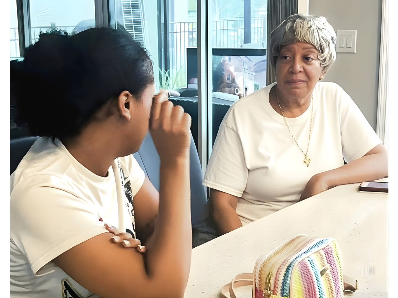 An older woman and a younger woman sit across from each other at a table, engaged in conversation, with glass doors in the background.