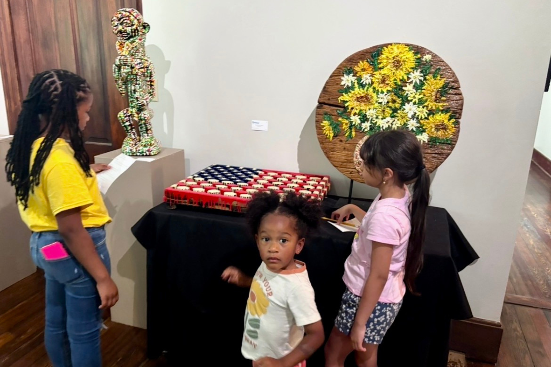 A group of children at an art exhibit. They are looking at, including a colorful sculpture of a dog and a round wooden table with a sunflower painting. The children are standing and looking at the art pieces, with one girl taking notes or sketching.