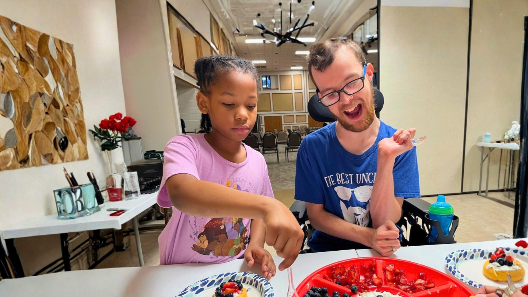 A young girl and a man are sitting at a table during a celebration, possibly a birthday party, with plates of sliced strawberries, blueberries, and a cake with berries. The girl appears focused on the food, while the man is smiling and holding a spoon, wearing glasses and a blue shirt. The background shows chairs, a decorative wall, and a black star-shaped light fixture.