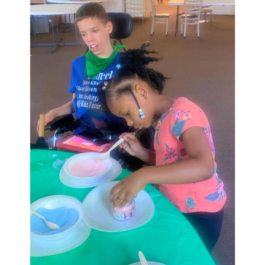 Children participate in a hands-on sensory activity designed to encourage creativity and connection. The table has other containers of colorful sand and utensils, with a green tablecloth, in a room with chairs and tables in the background.