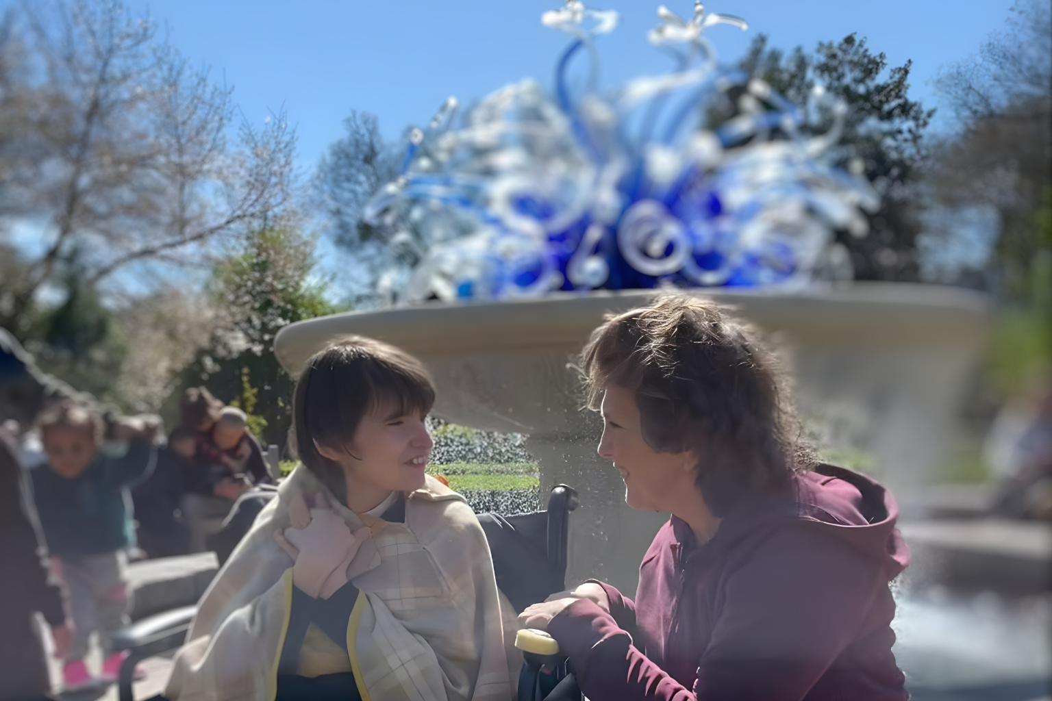 Mother and daughter are enjoying an outdoor community experience, focused on connection and shared experience. Background: a large decorative glass sculpture outdoors with trees and a blue sky in the background.