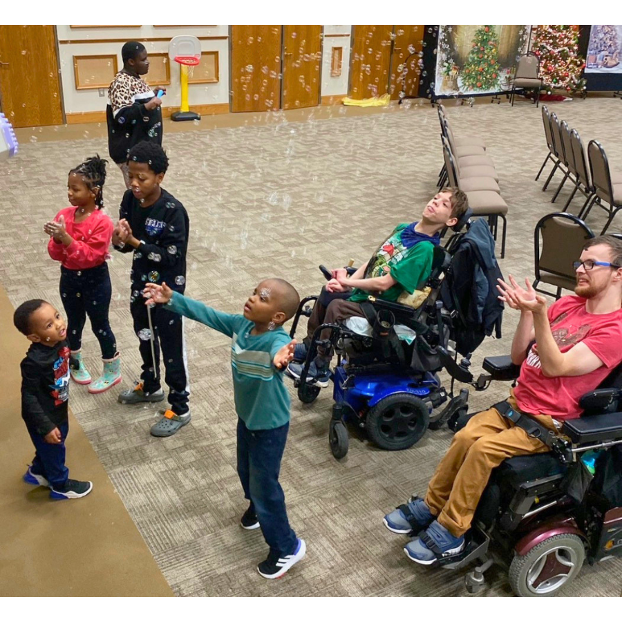 Children of all abilities are enjoying an inclusive activity together; they are playing with bubbles, with one blowing bubbles and others expressing curiosity or joy. Two children are in wheelchairs, and all are observing the bubbles.