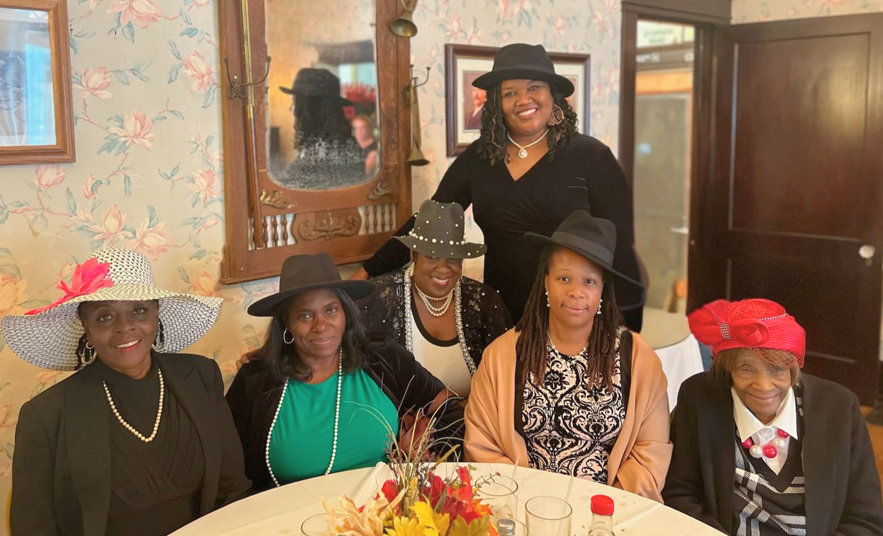 Six women wearing fancy hats and pearls sit and stand around a decorated table in a room with floral wallpaper and framed pictures. One woman stands behind the others, smiling.