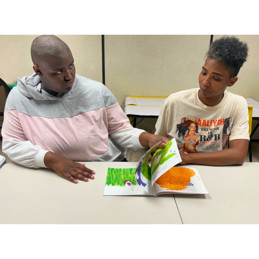 Youth and adult volunteers engaged in a shared reading and educational activity in a supportive learning setting at a table, looking at a colorful children's book with illustrations of animals and a large orange object.
