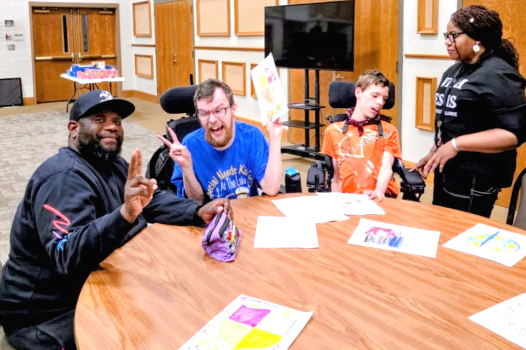 A group of four people sitting around a wooden table in a room. They are smiling and gesturing, with some holding papers and coloring pages on the table.