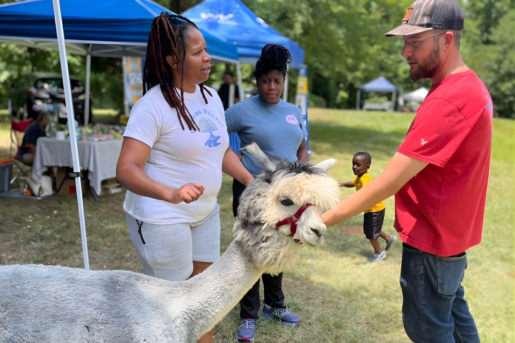 Families and children are participating in a fun outdoor community engagement event together. Some are interacting with a small white llama with black markings on its face, standing on green grass under trees, with tents and tables in the background.
