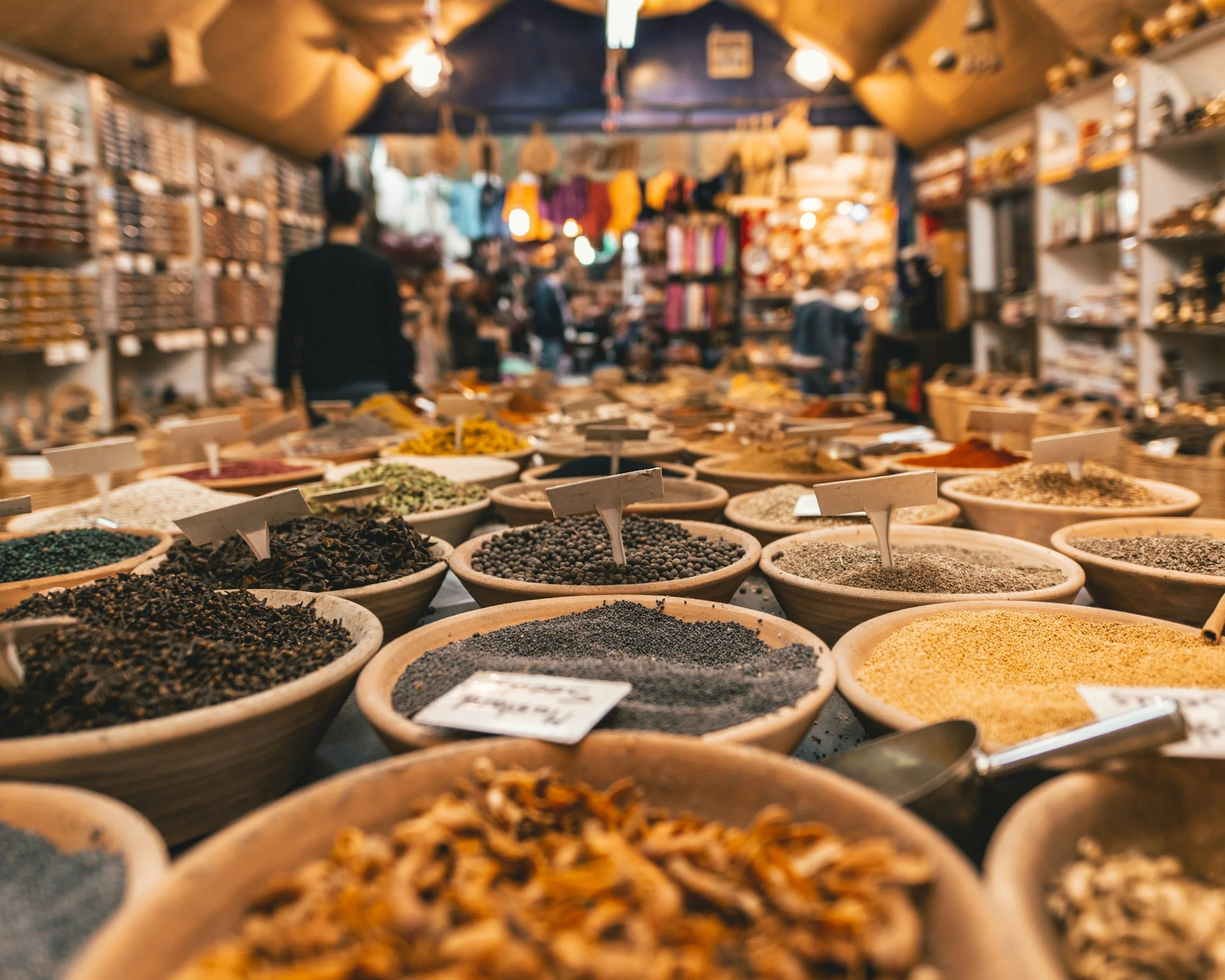 A market stall displaying numerous bowls of various spices and herbs with small labels, indoor setting with shelves filled with products and shoppers shopping in the background.