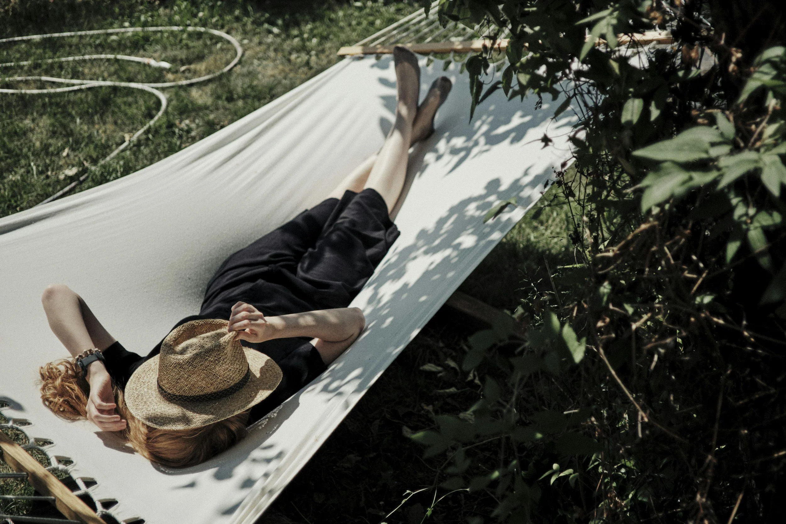 Person lying on a white fabric slide outdoors, wearing black clothing and a large straw hat, with a jungle or dense foliage in the background.