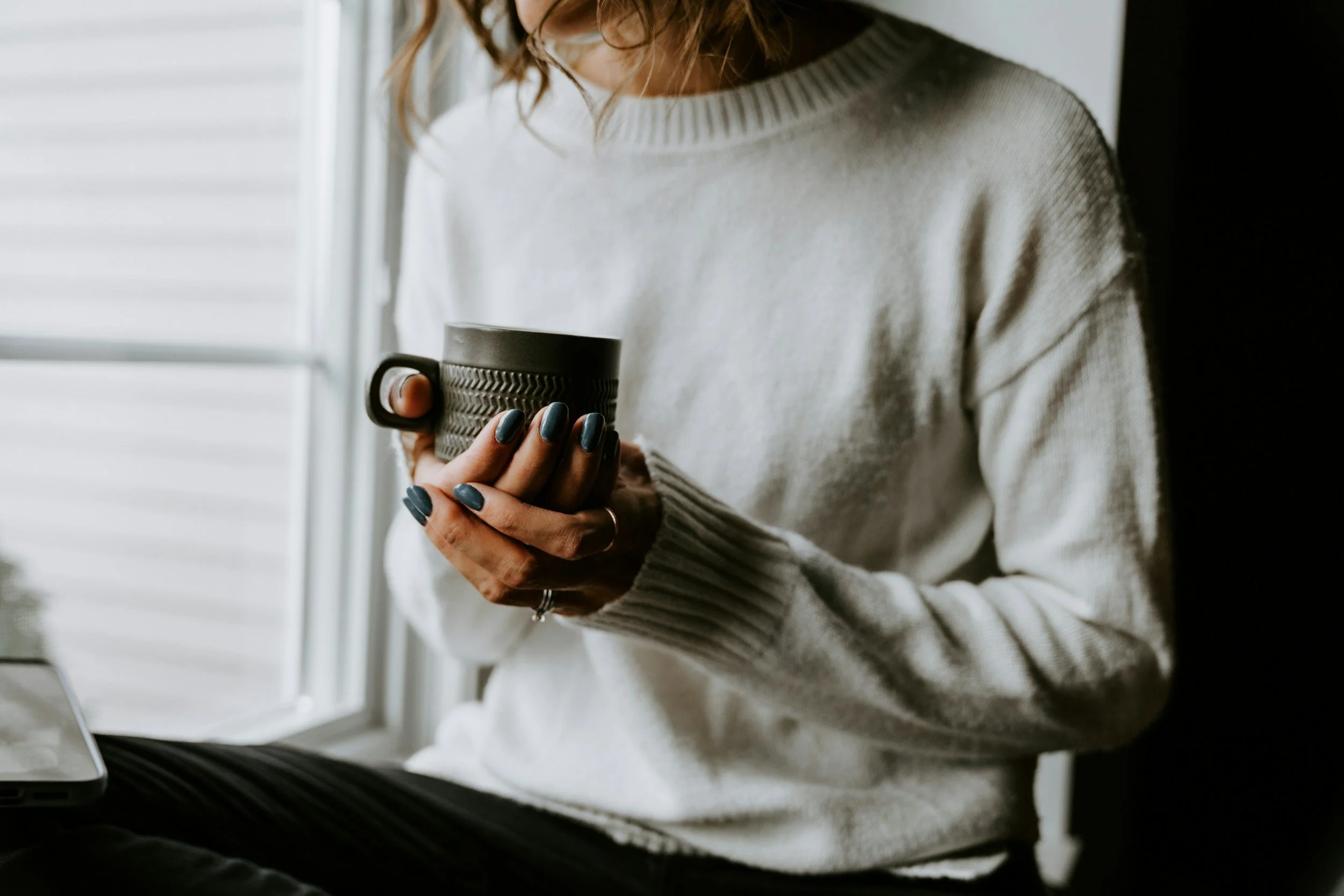 A woman in a light gray sweater holding a dark gray coffee mug while sitting near a window.