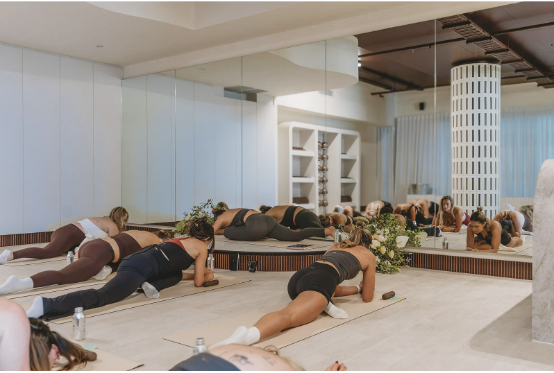 Group of women participating in a yoga or fitness class in a spacious, modern studio with large mirror, floral arrangement, and minimalist decor.