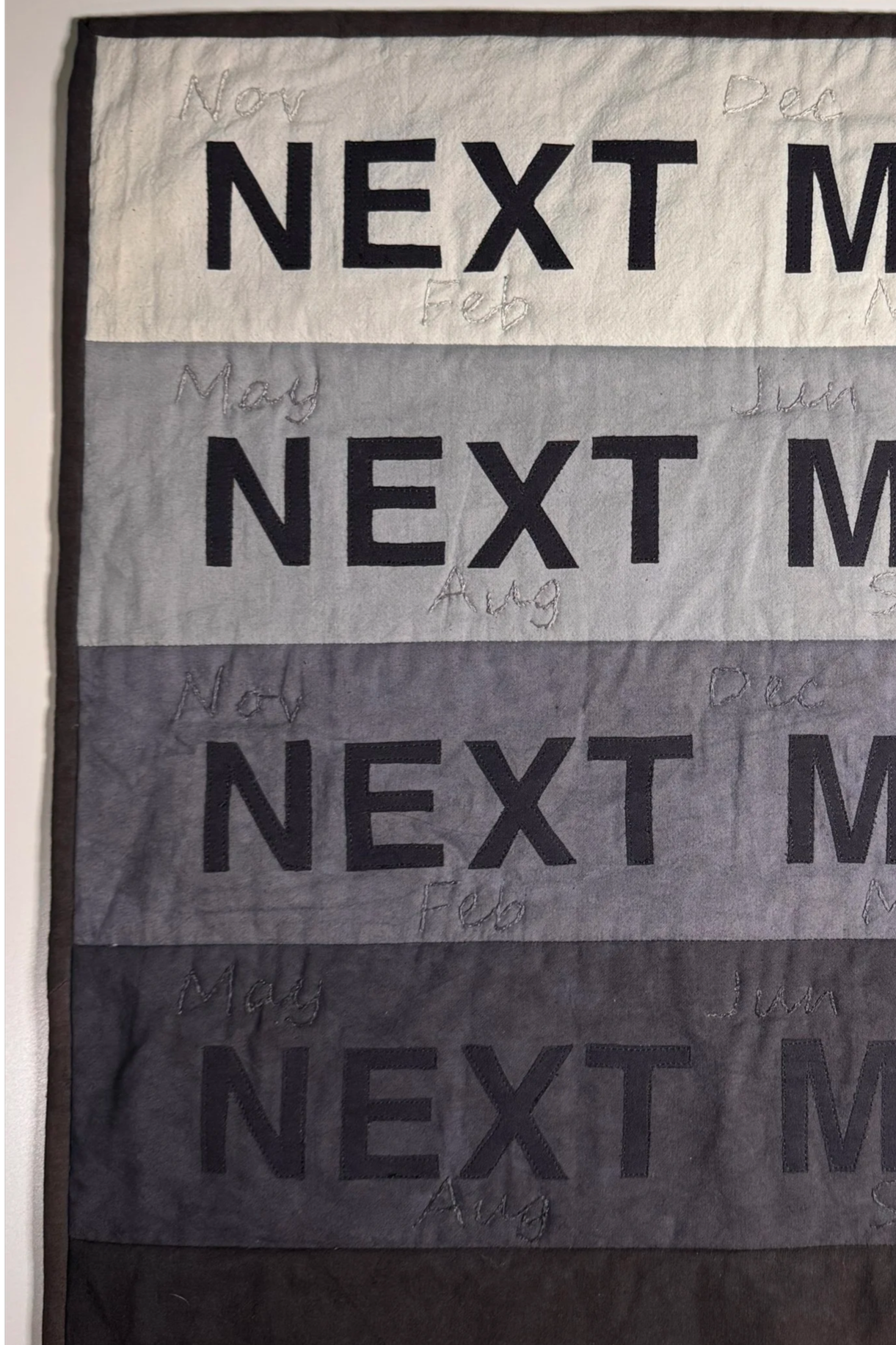 A fabric or quilt with four horizontal sections, each showing the phrase "NEXT MONTH" in large black letters and the months from October to December stitched in handwritten script, indicating a countdown or anticipation for the upcoming months.