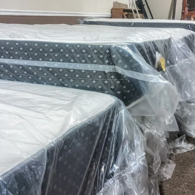 Multiple mattresses wrapped in clear plastic in a storage or warehouse setting.
