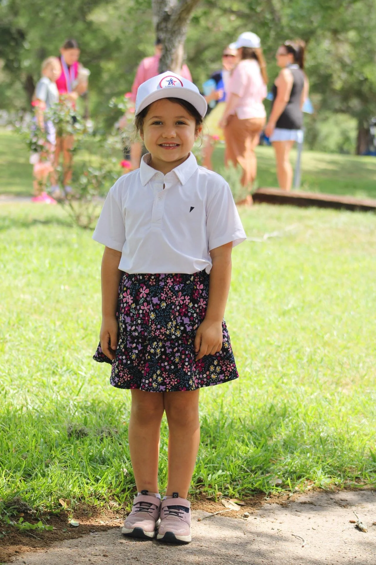 Smiling young girl in white shirt, floral skirt, pink sneakers, wearing white hat, standing outdoors on a grassy area with trees and a group of people in the background