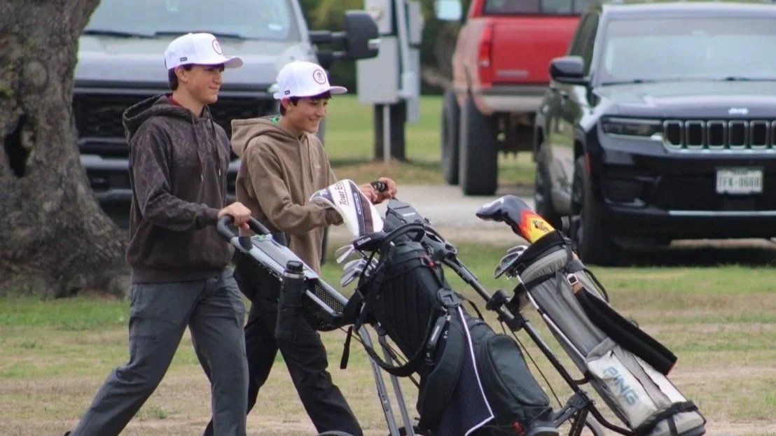 Two young males walking on grass, each pushing a golf cart loaded with golf bags, wearing caps and casual clothing.