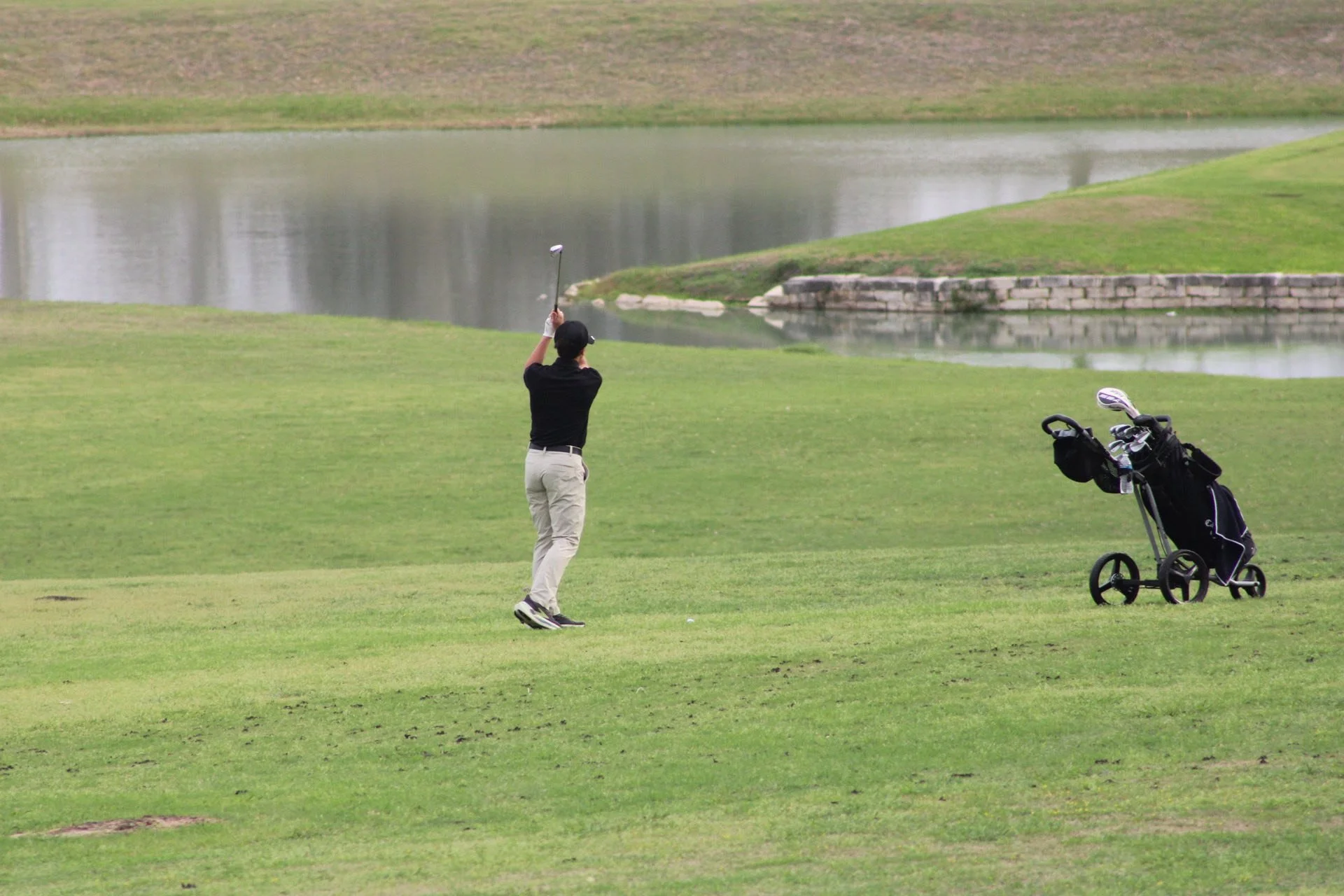 A person playing golf near a water hazard with a golf cart and bag on a golf course.