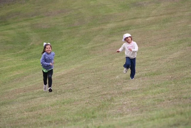 Two children, a girl and a boy, running on a grassy hill, smiling and enjoying the outdoor activity.