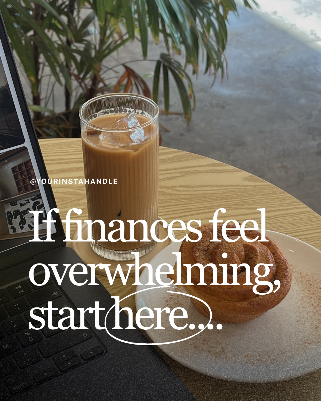 A laptop, a glass of iced coffee, and a cinnamon roll on a white plate on a wooden table with a green plant in the background.