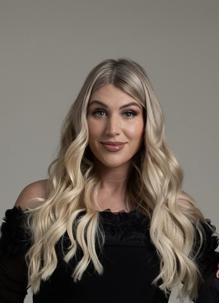 A woman with long, wavy blonde hair, wearing a black off-the-shoulder top, smiling and looking at the camera against a plain gray background.