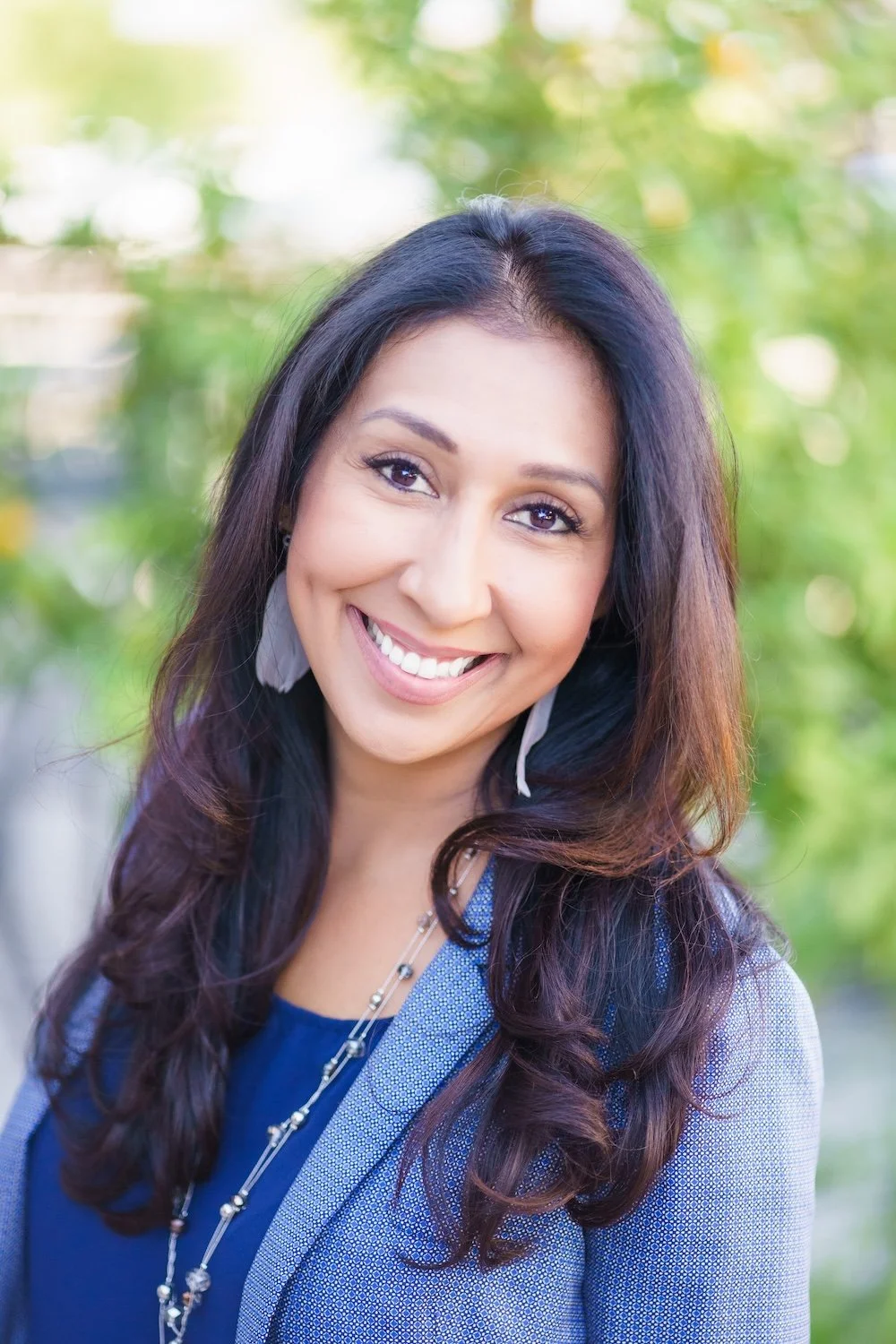 A woman with long dark hair smiling outdoors with a blurred green background.