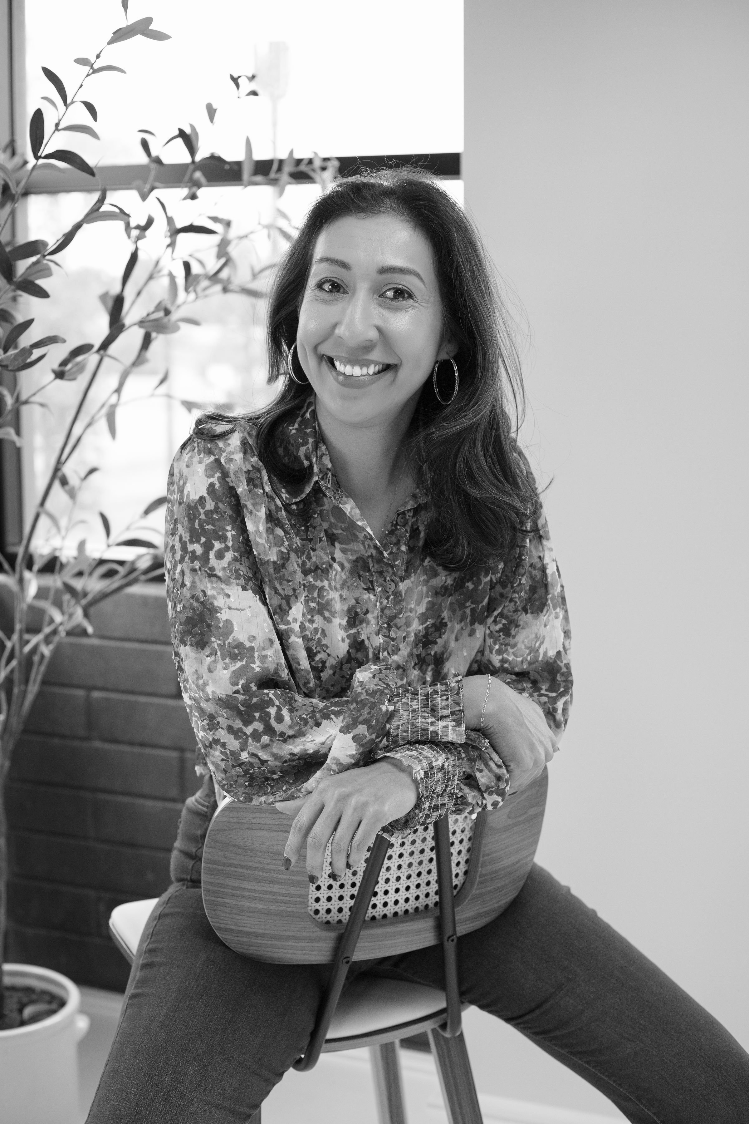 A woman with shoulder-length hair, wearing hoop earrings and a patterned blouse, smiling while sitting on a chair, with a potted plant and a window in the background.