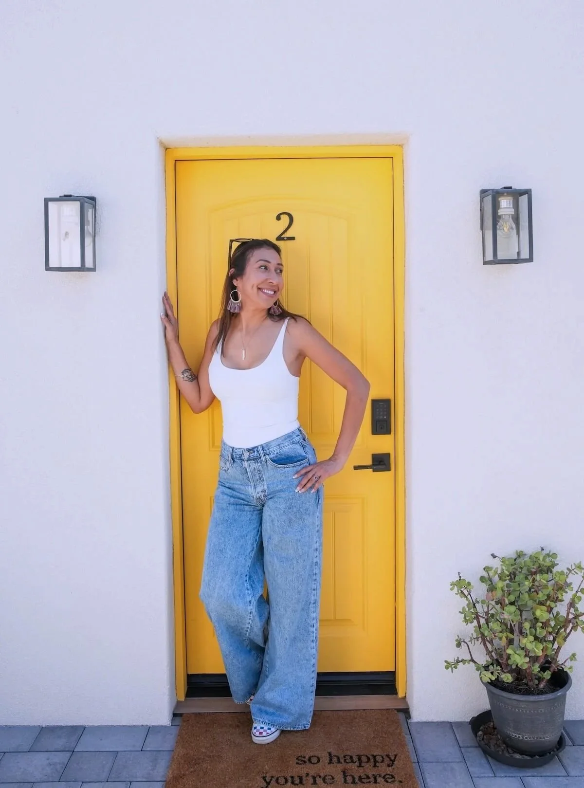 Woman standing at a yellow front door, smiling, wearing a white tank top and blue jeans, with a plant to the right.