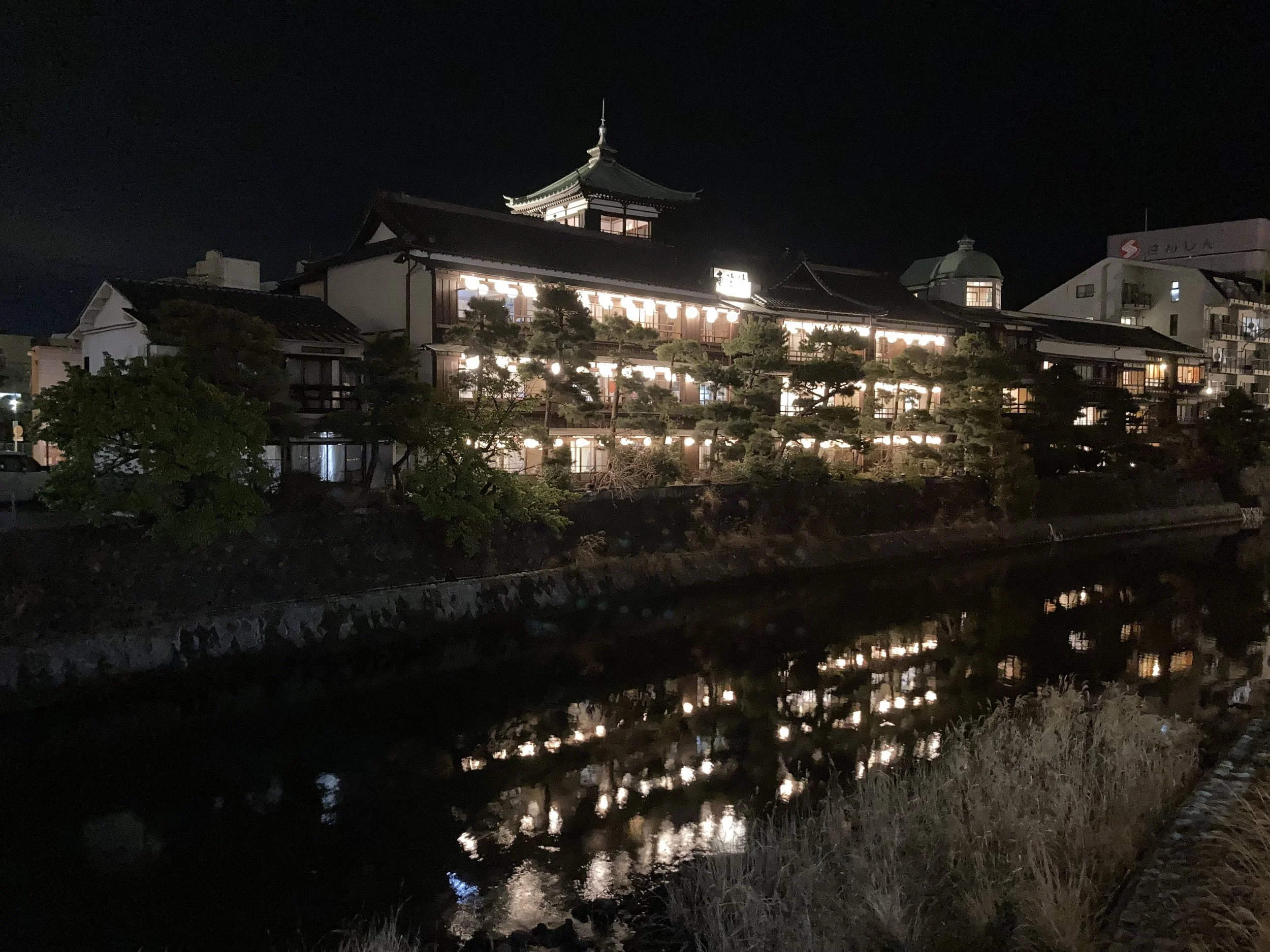 Night view of a traditional Japanese building with multiple floors, illuminated windows, and a pagoda-style roof, reflected in a river in the foreground.