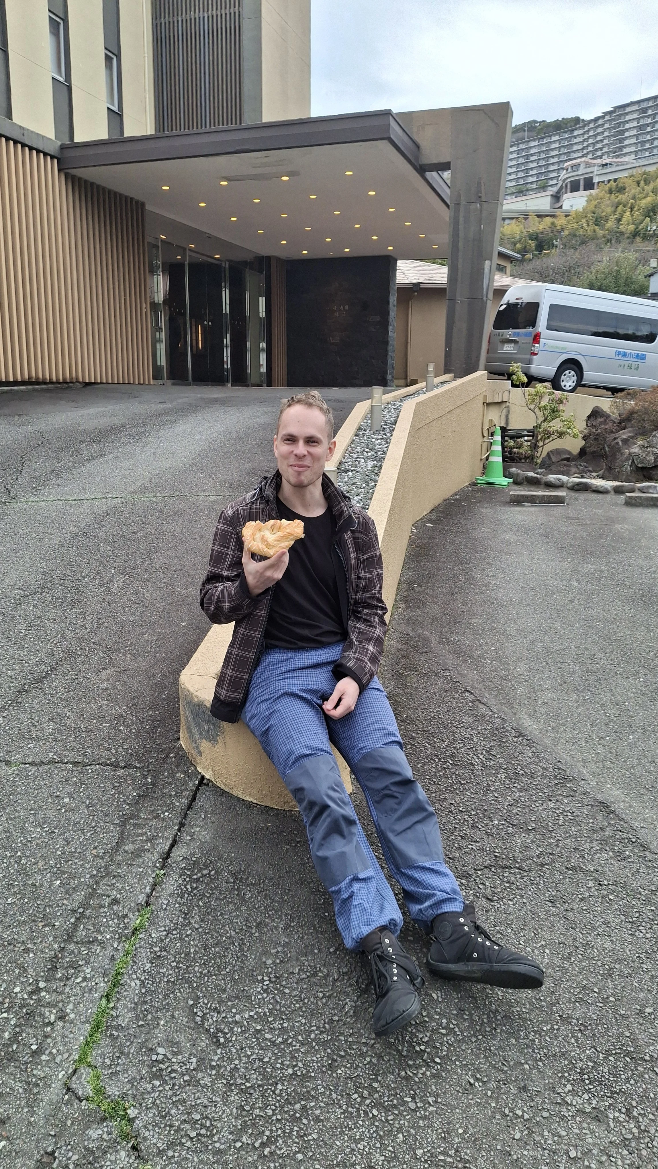 Ondřej, one of the guides, sitting on a curved concrete bench outside a modern building, holding a pastry and smiling.