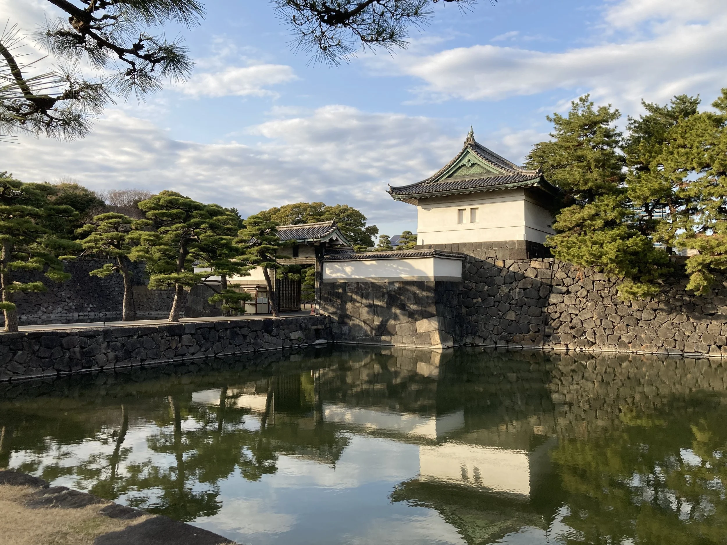 A traditional Japanese building with a pond in the foreground, surrounded by neatly trimmed trees and stone walls, under a partly cloudy sky.