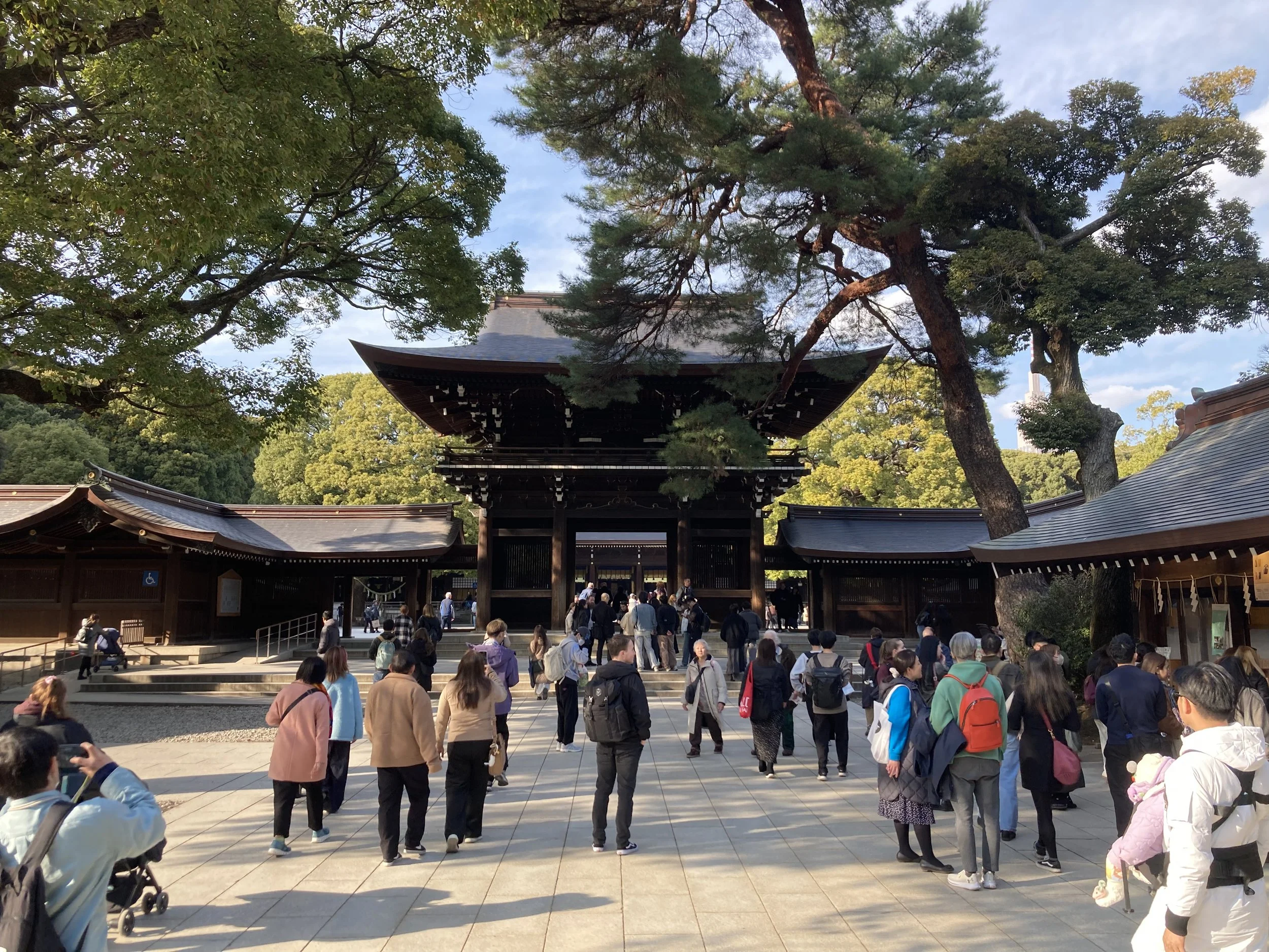 People gathered in front of a traditional Japanese wooden gate surrounded by trees in a park or temple grounds.