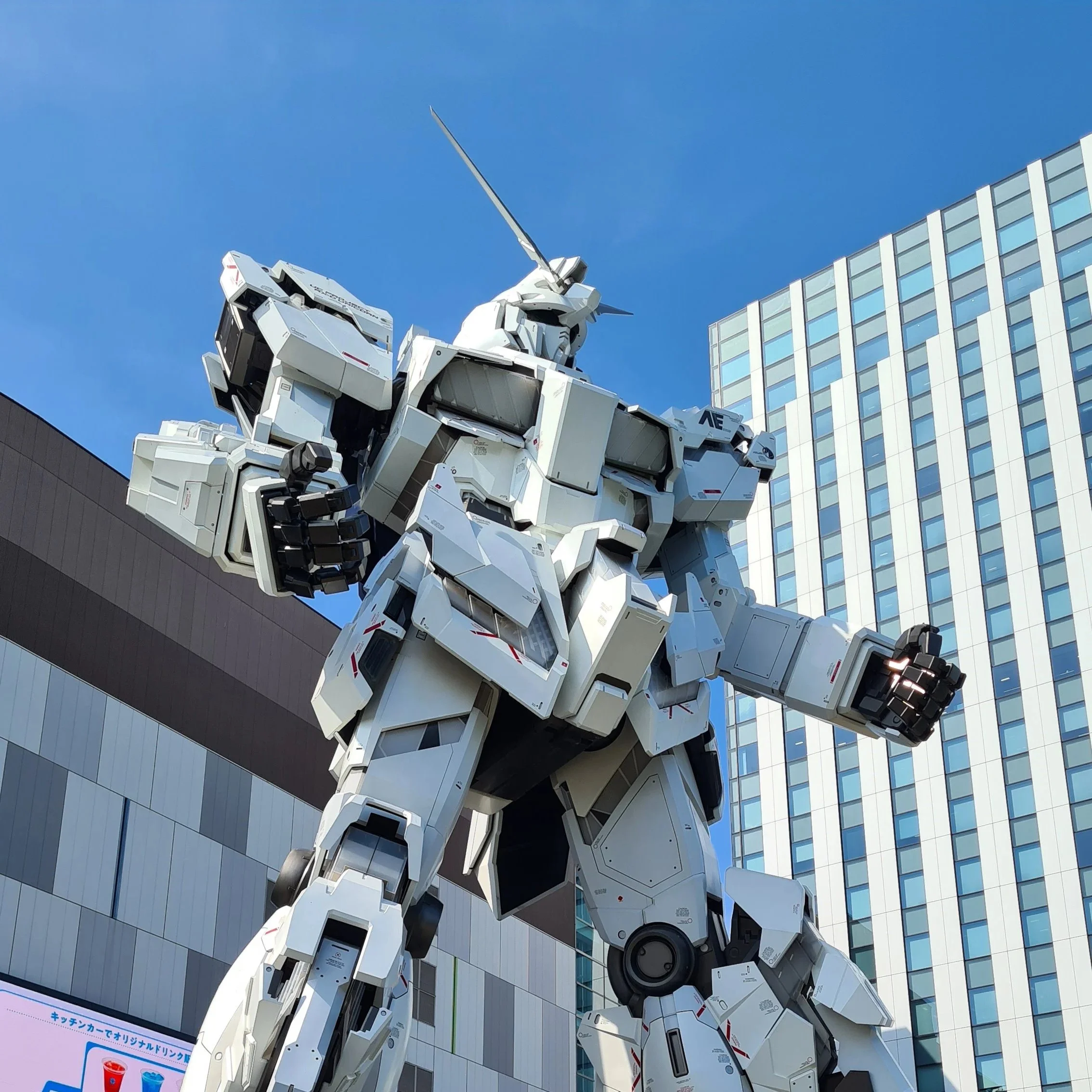 Large white Gundam robot sculpture outdoors against a backdrop of modern high-rise buildings and a clear blue sky.
